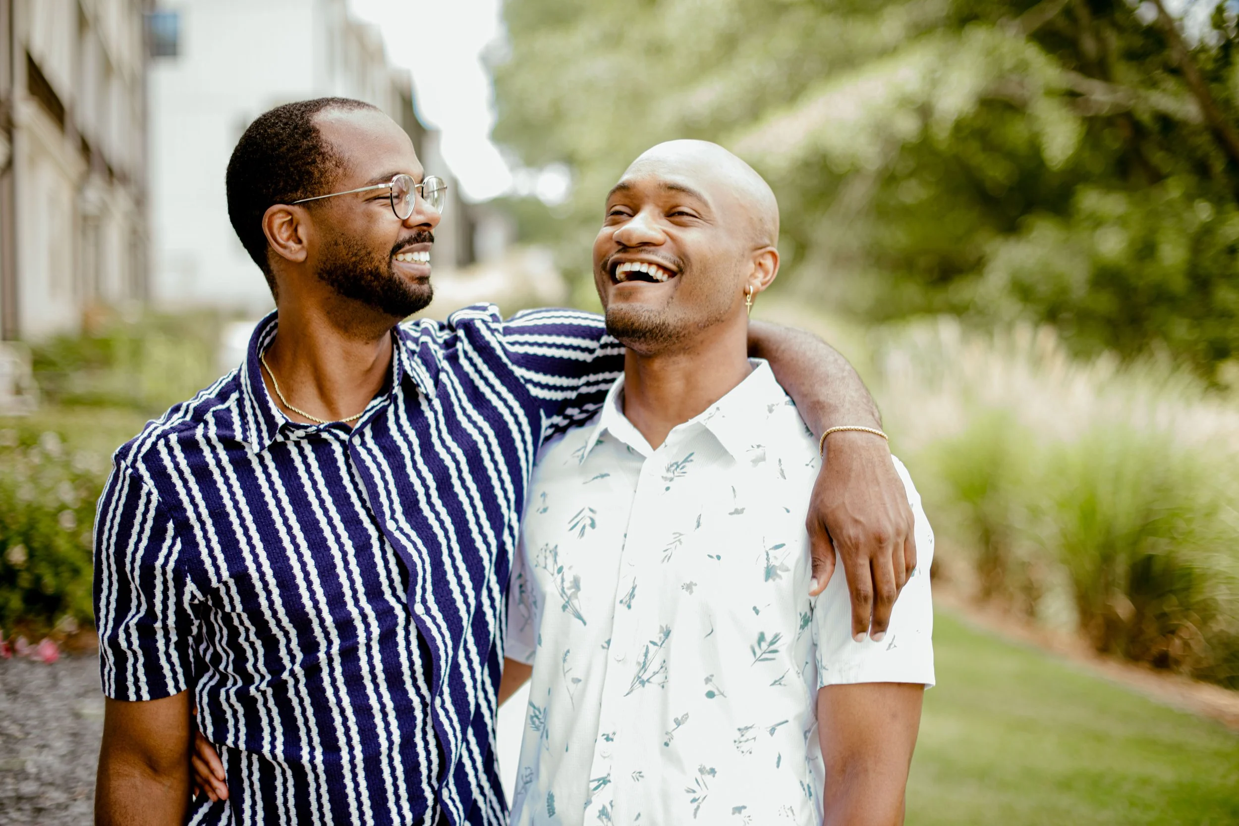 Two men smiling and embracing outdoors with trees and buildings in the background.