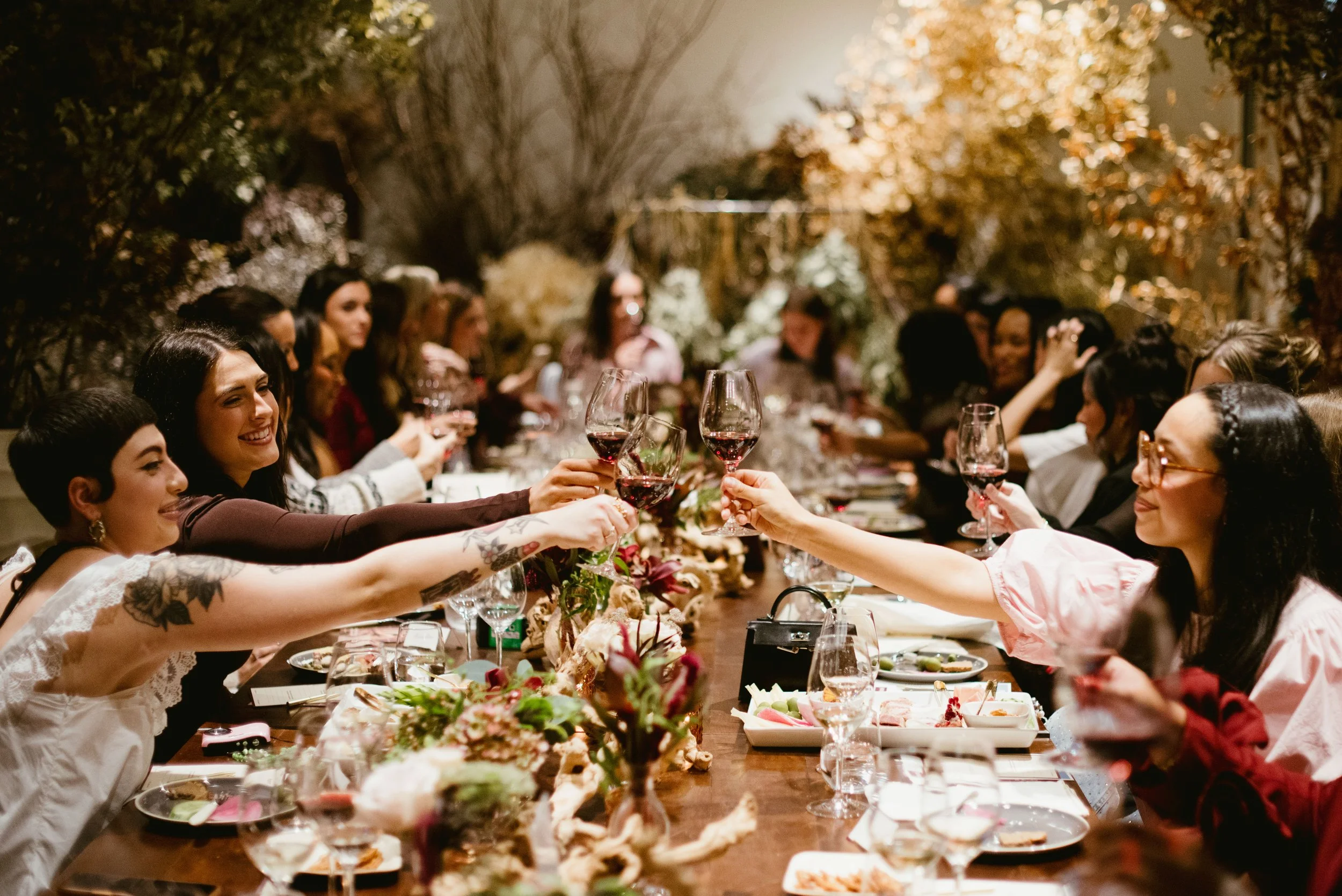 People at a long dinner table raising wine glasses in a toast. The setting includes floral centerpieces and a warm, cozy ambiance with many women and some men smiling and enjoying the gathering.
