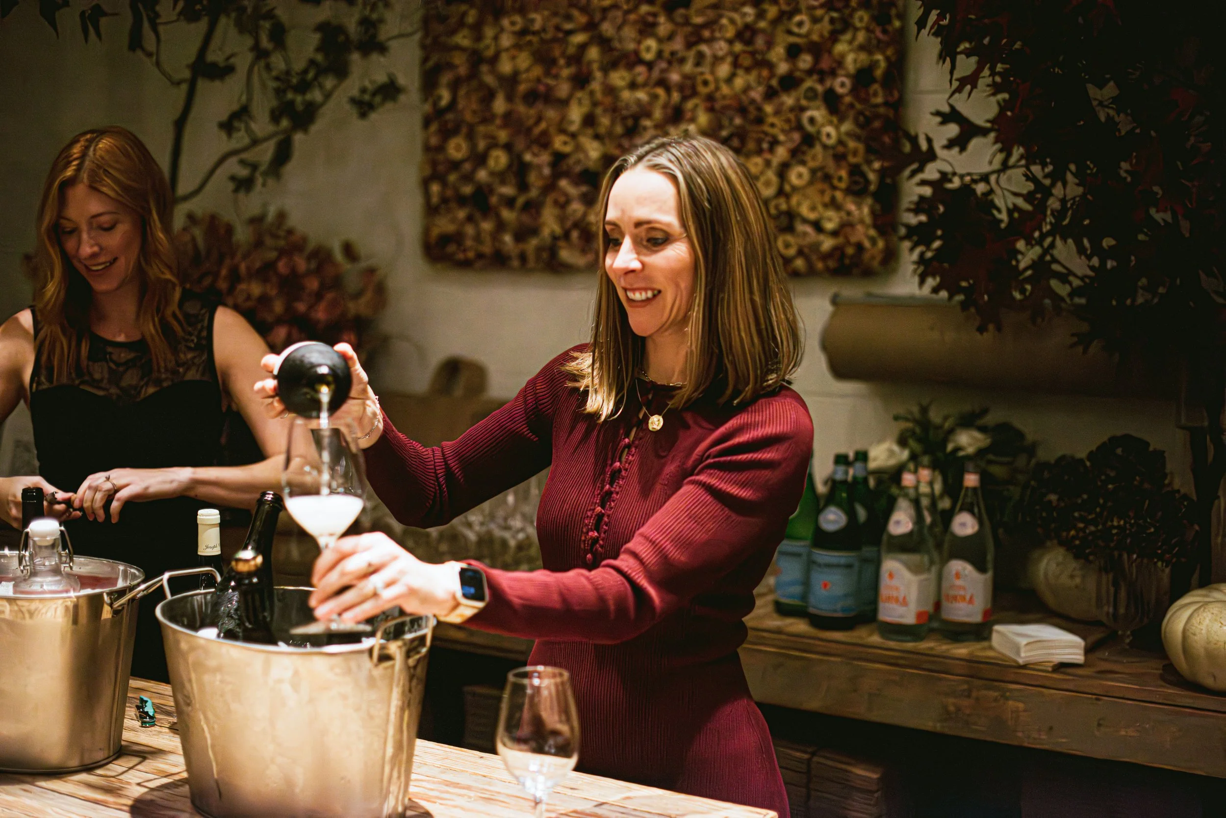 Two women at a bar or restaurant, preparing drinks. One woman in a red dress is pouring a liquid into a glass, while the other woman in a black top is smiling and looking down. The background features shelves with bottles and decorative wall art.