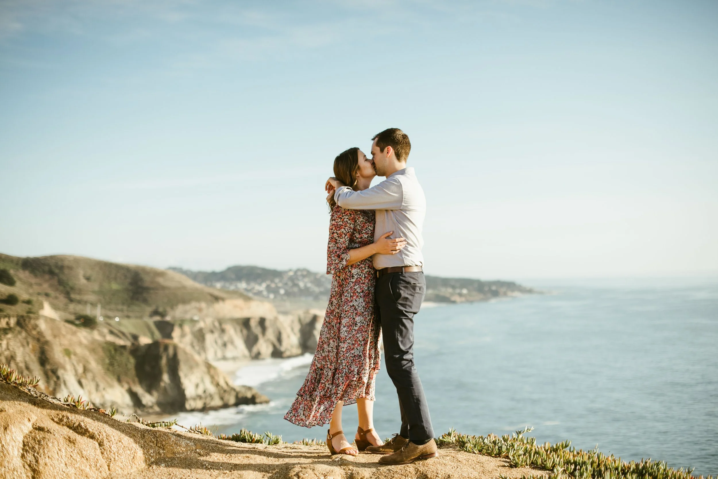 A couple stands on a cliffside kissing with the ocean and cliffs in the background during daytime.