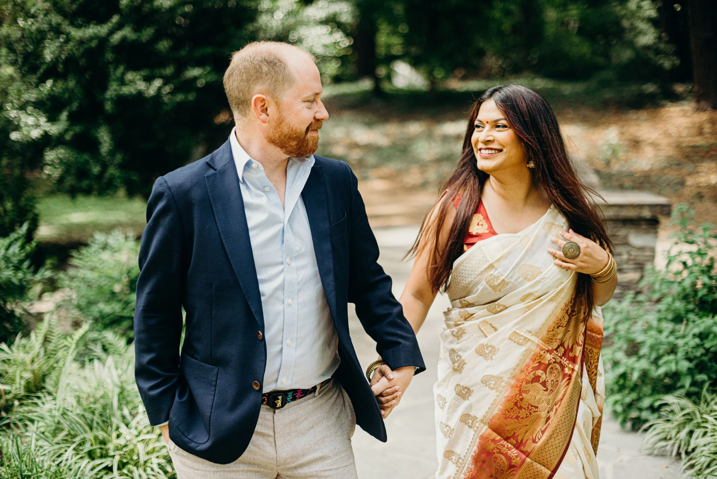 A man and woman holding hands and smiling at each other while walking outdoors in a park surrounded by greenery.
