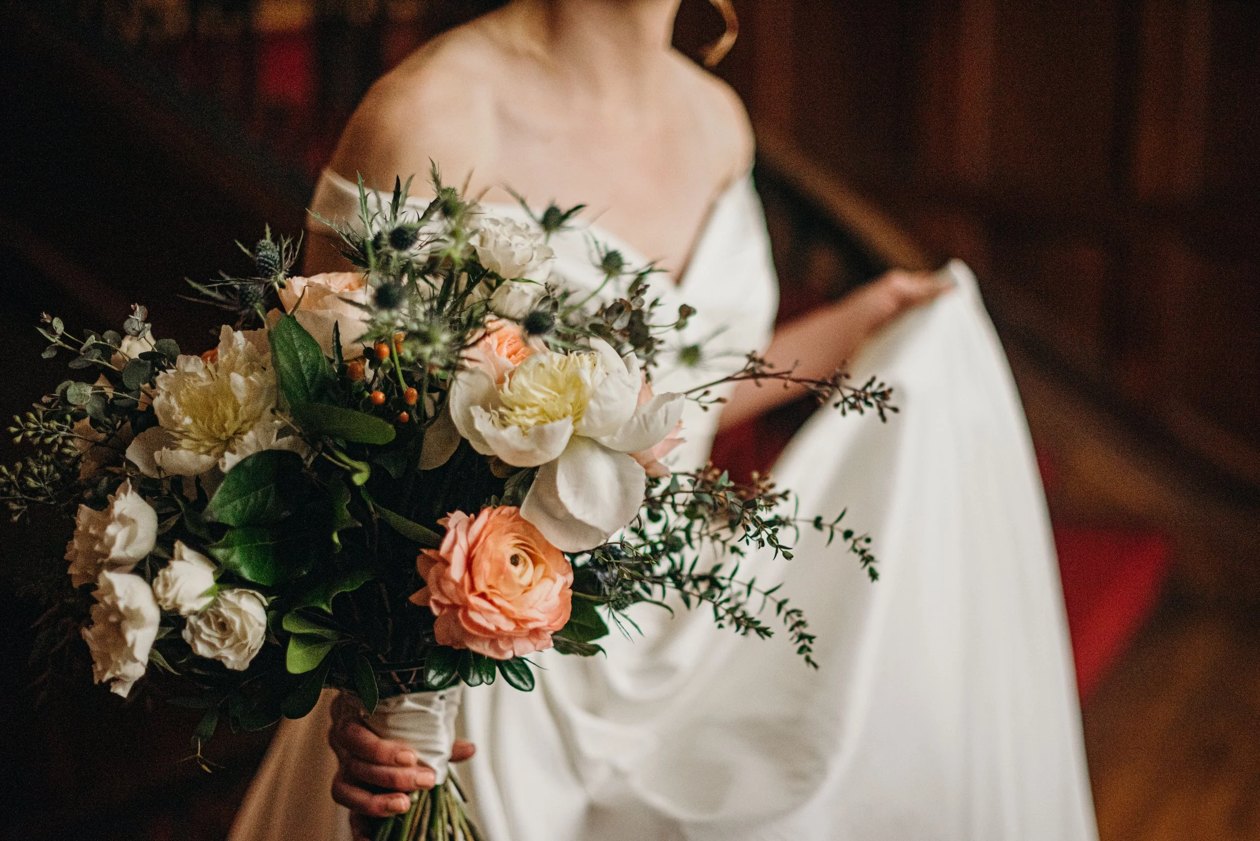 A bride holding a bouquet of flowers, dressed in an off-the-shoulder wedding gown with a blurred background.