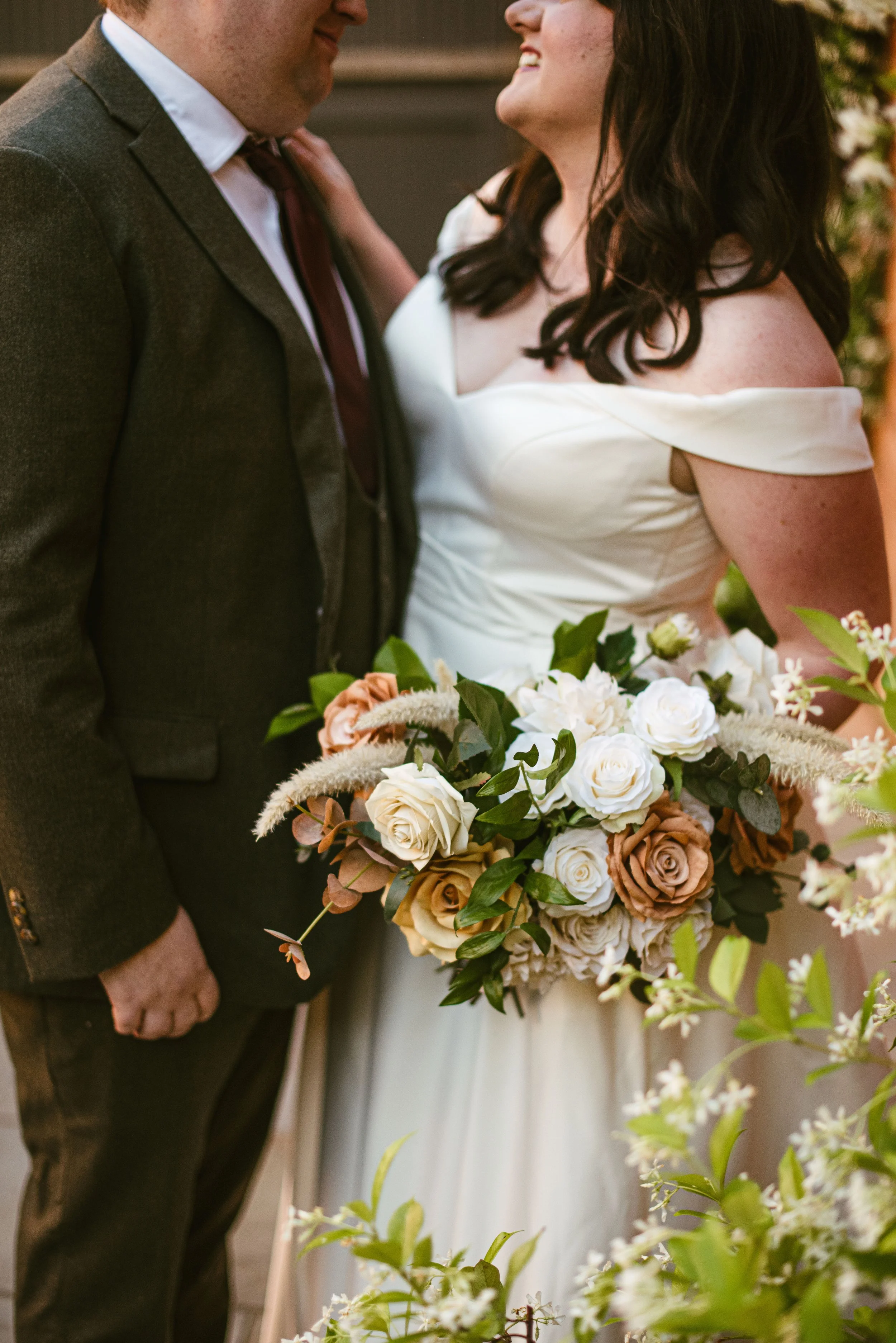 A close-up of a couple at a wedding, focusing on their faces and upper bodies. The woman has dark hair and is wearing a white off-the-shoulder wedding dress, holding a large bouquet of white and brown roses with greenery. The man is dressed in a dark