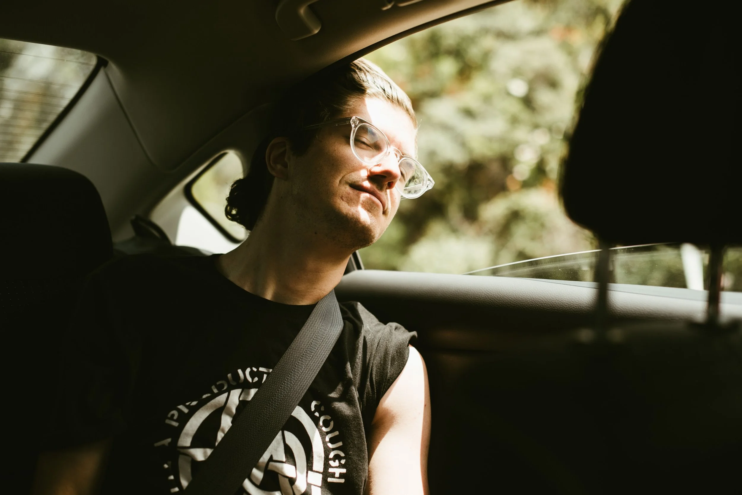 A young man wearing glasses and a black t-shirt, smiling with eyes closed, sitting in the back seat of a car, with trees visible outside the window.