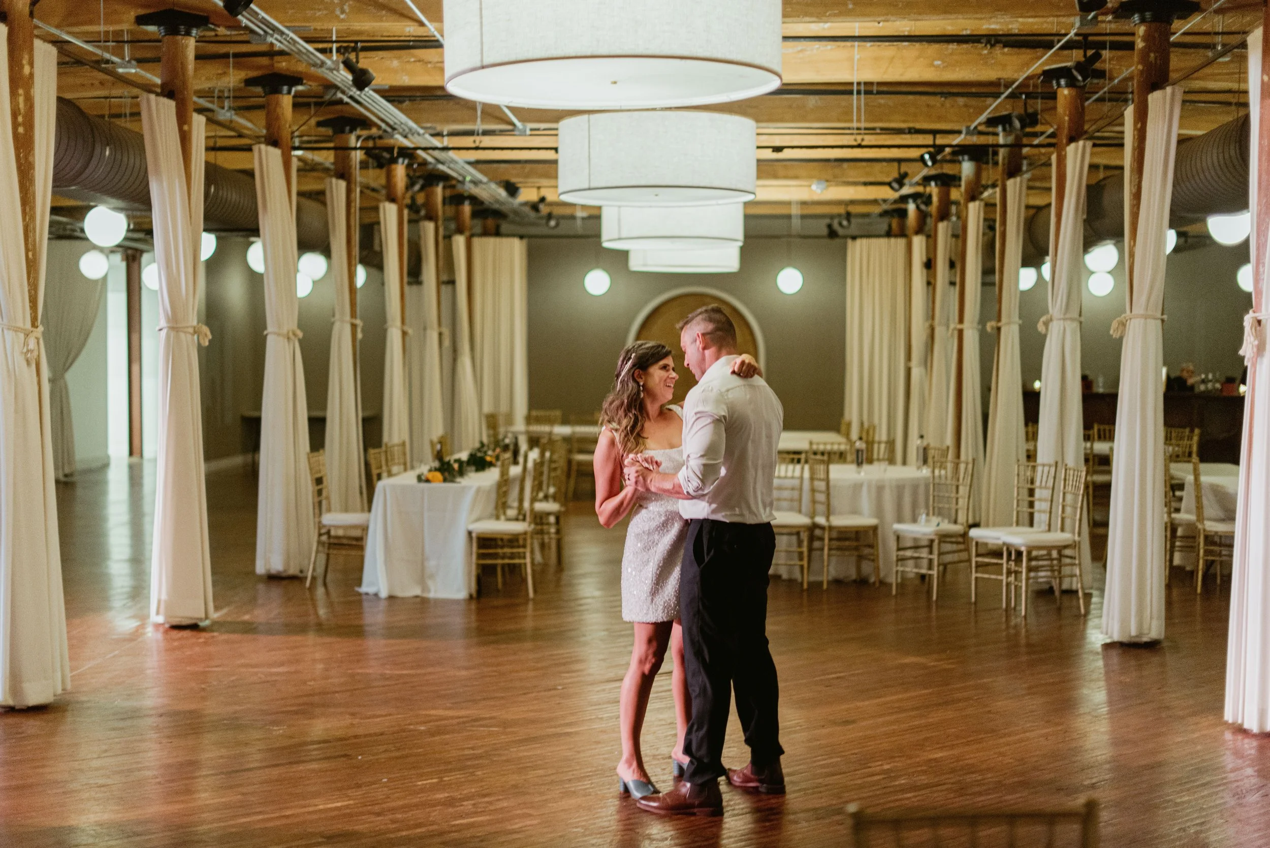 A couple dances during their wedding reception in a decorated banquet hall with round tables, chairs, and large hanging lights.