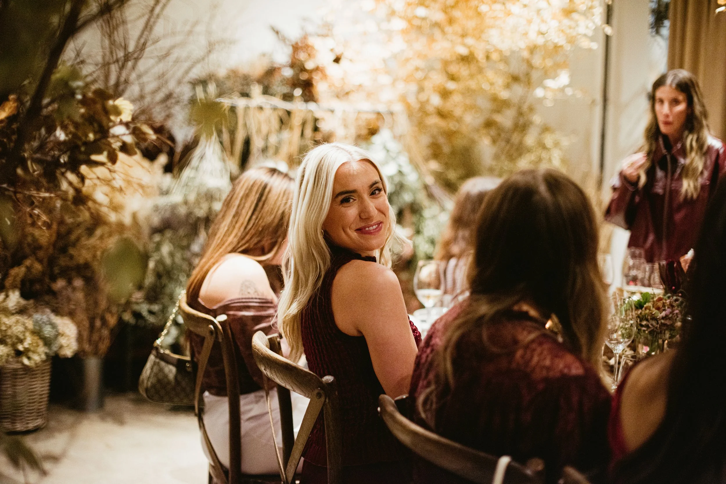 A group of women sitting at a dinner table decorated with flowers. One woman with long blonde hair and a smile is looking at the camera. The setting has warm lighting and a festive ambiance.