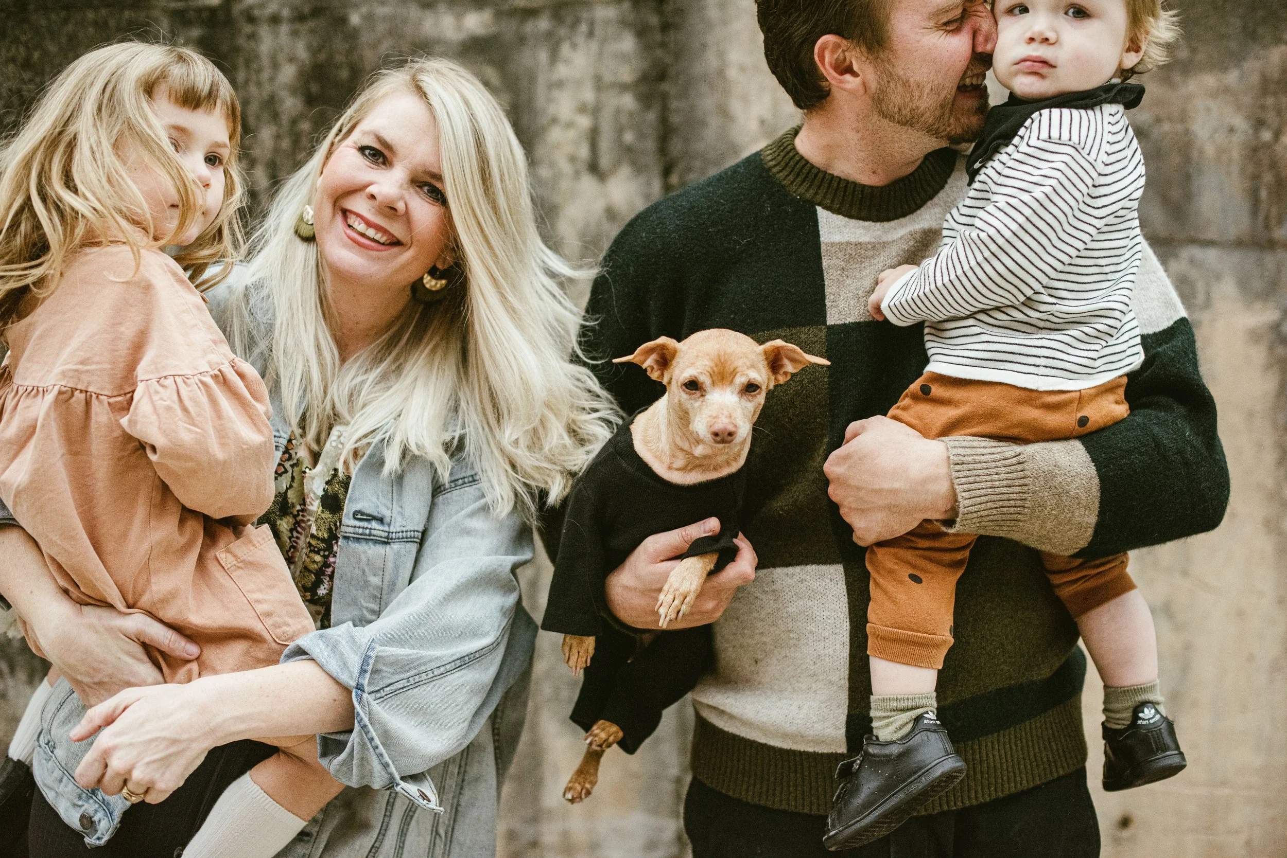 A family of four with a dog, standing outdoors near a brick wall. The mother with long blonde hair and a denim jacket is hugging a young girl with long wavy blonde hair, wearing a peach-colored dress. The father with a beard is holding a small boy wi