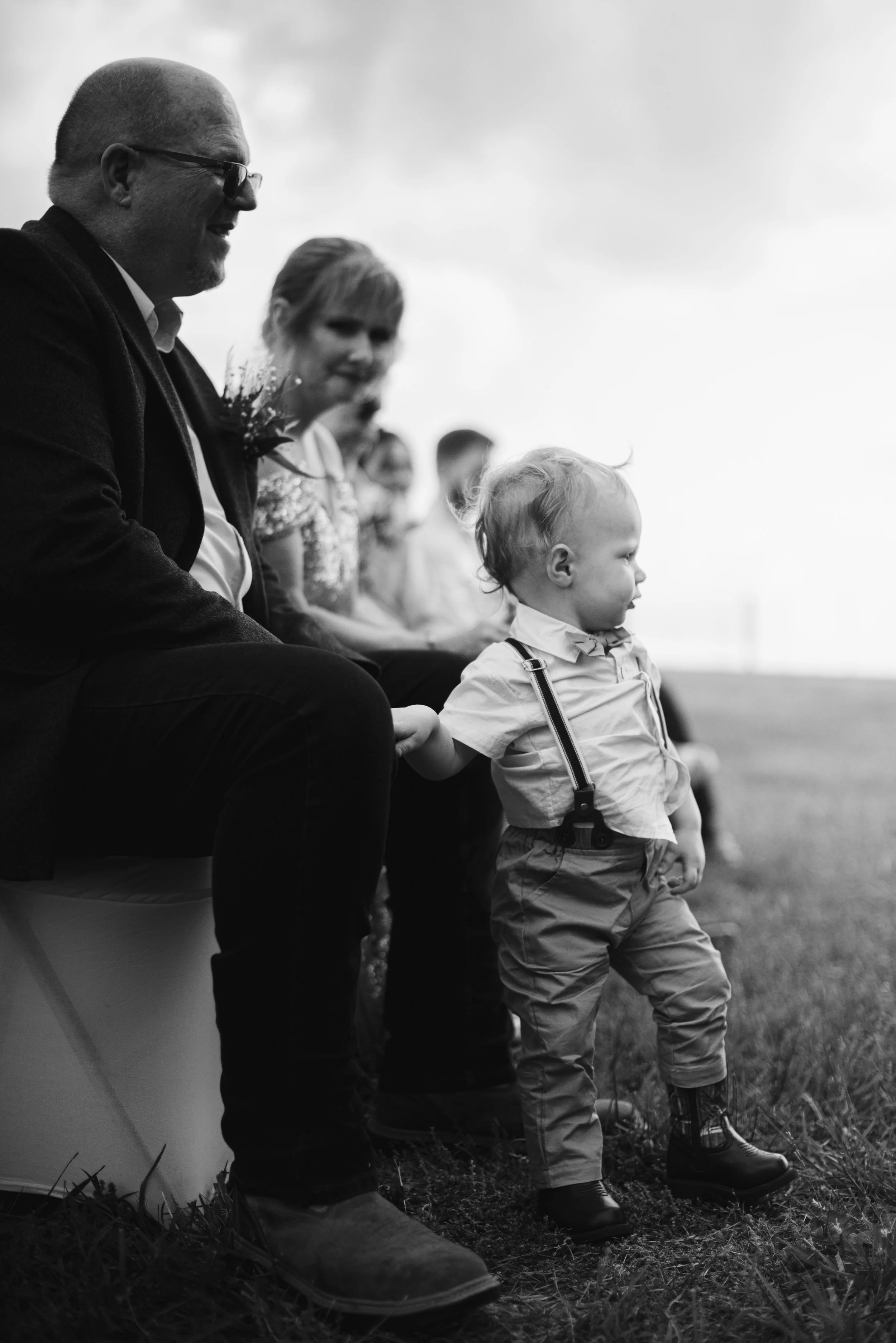 Black and white image of a man, woman, and young boy sitting outdoors on a grassy area, facing to the right. The man is wearing glasses and a jacket, the woman has a flower pinned to her clothing, and the boy is dressed in a shirt, suspenders, and bo