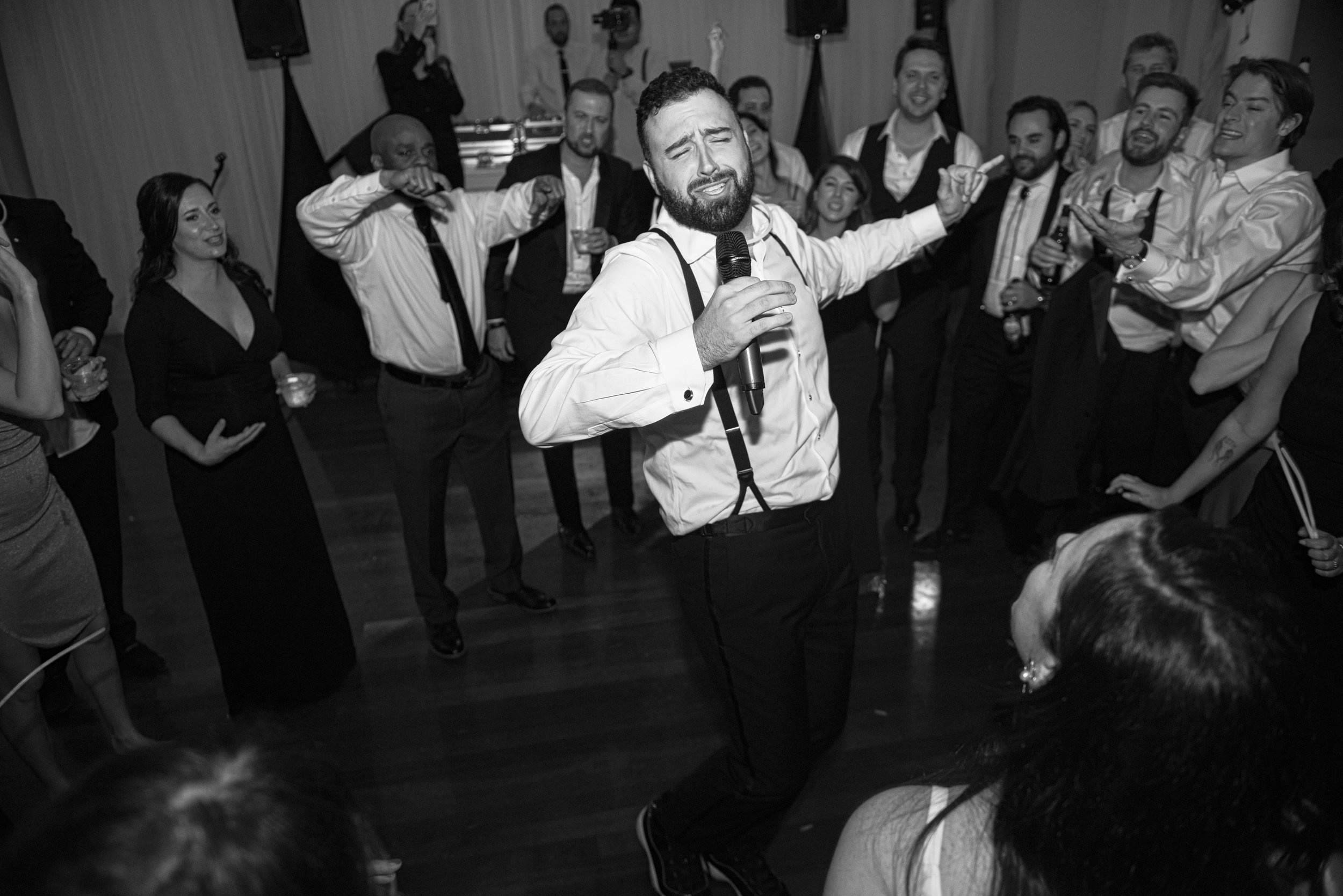 A man with a beard singing into a microphone at a social gathering with people dancing and smiling in the background.
