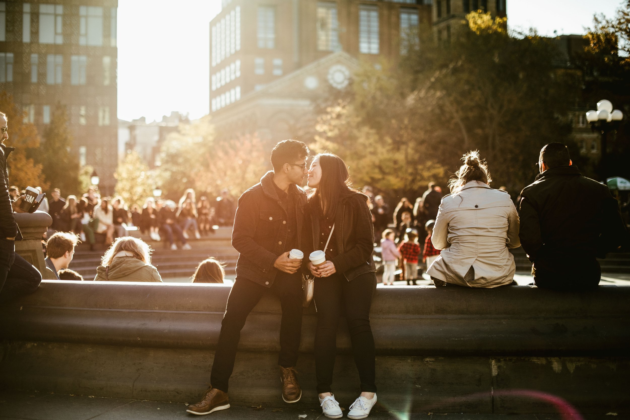 A couple kissing and holding coffee cups in a city park during sunset, surrounded by other people sitting and standing, with tall buildings and trees in the background.