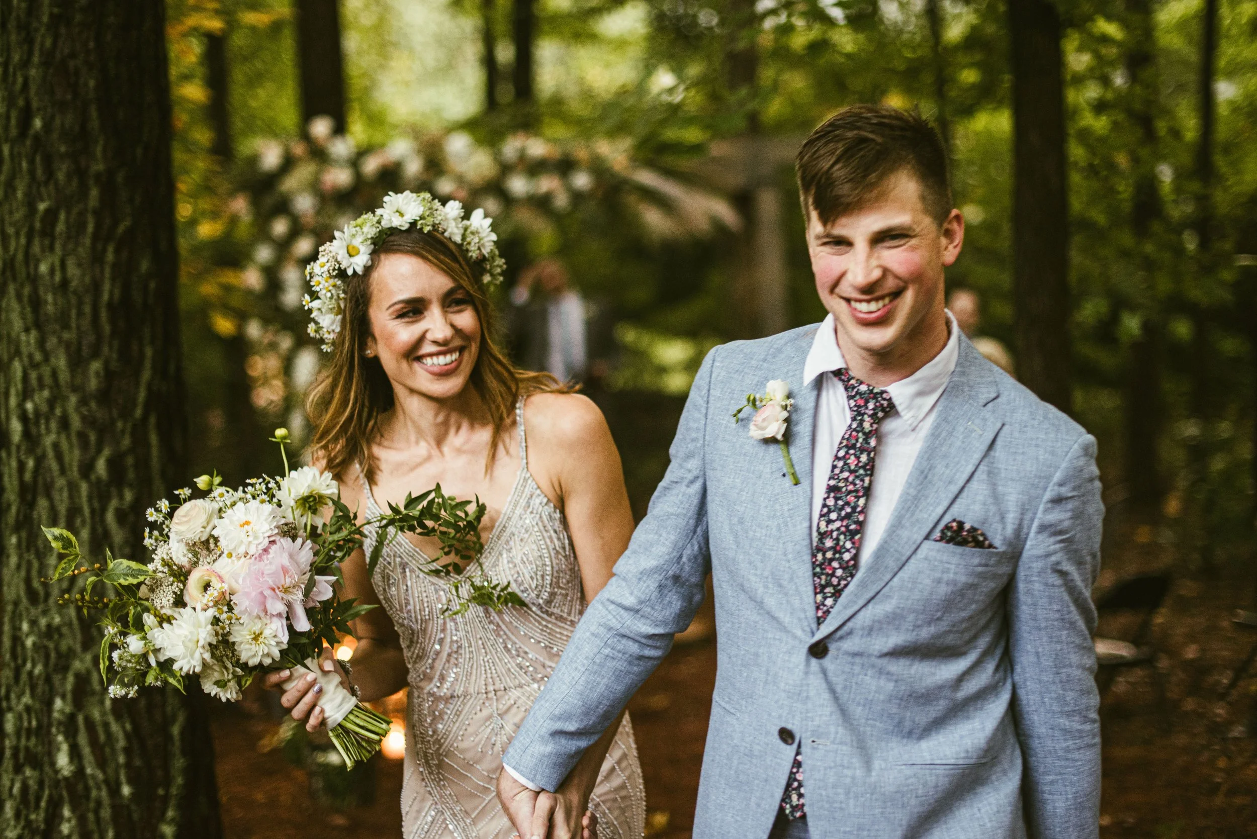 A happy couple walking hand in hand outdoors during their wedding ceremony in a wooded area. The bride is holding a large bouquet of flowers and wearing a floral crown, while the groom is dressed in a light blue suit with a floral tie and boutonnière