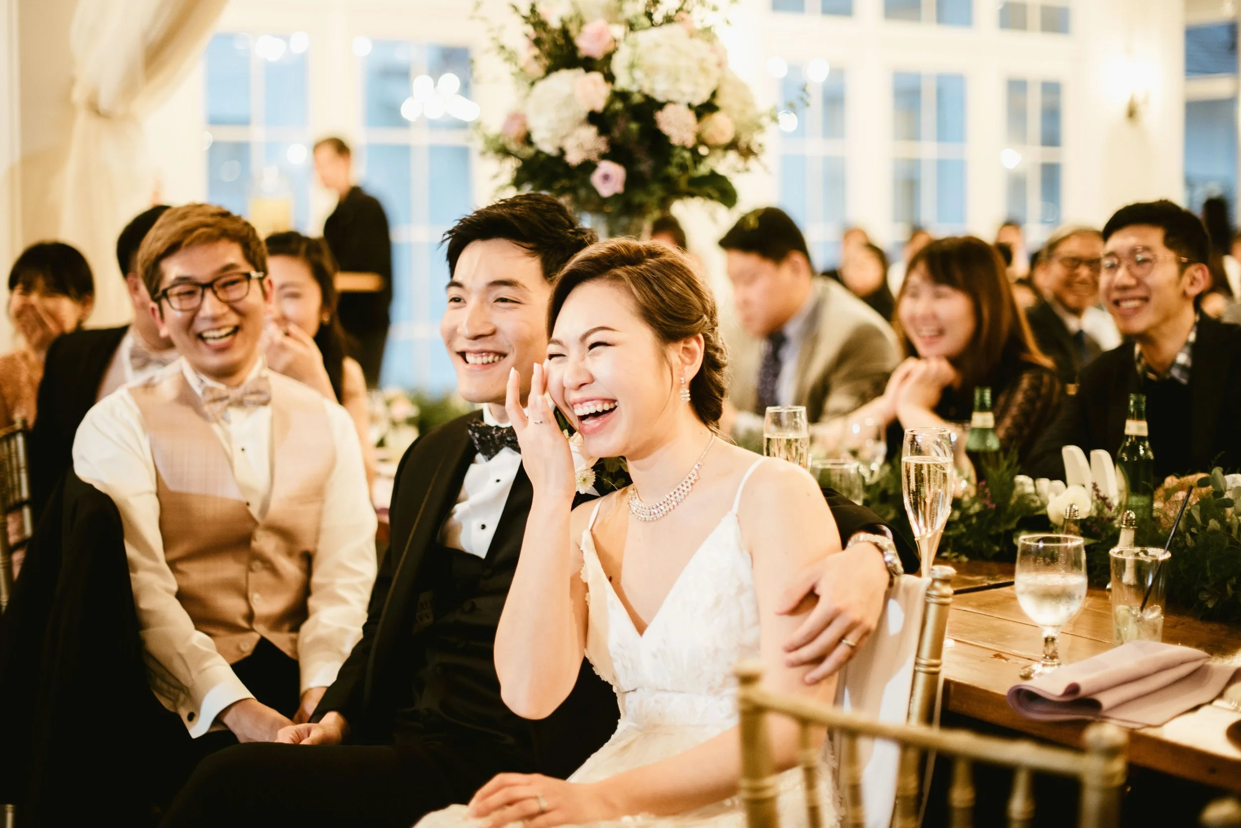 A group of people at a wedding reception, happily laughing and enjoying their time. The bride and groom are in the foreground, dressed in formal attire, with the bride in a white wedding dress and the groom in a tuxedo. The background shows a decorat