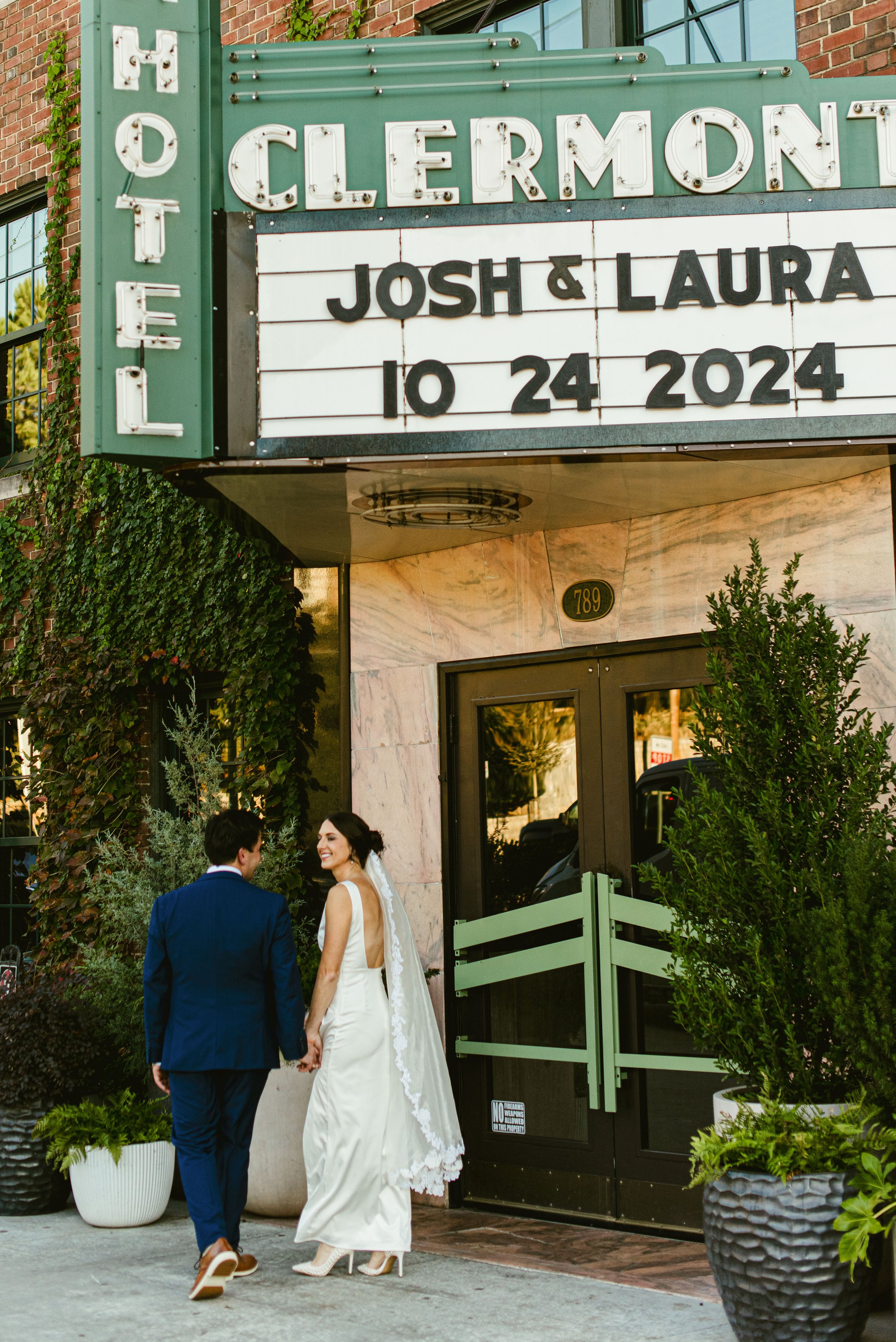 A bride and groom holding hands outside a wedding venue with a marquee sign, greenery in pots and a glass entrance.