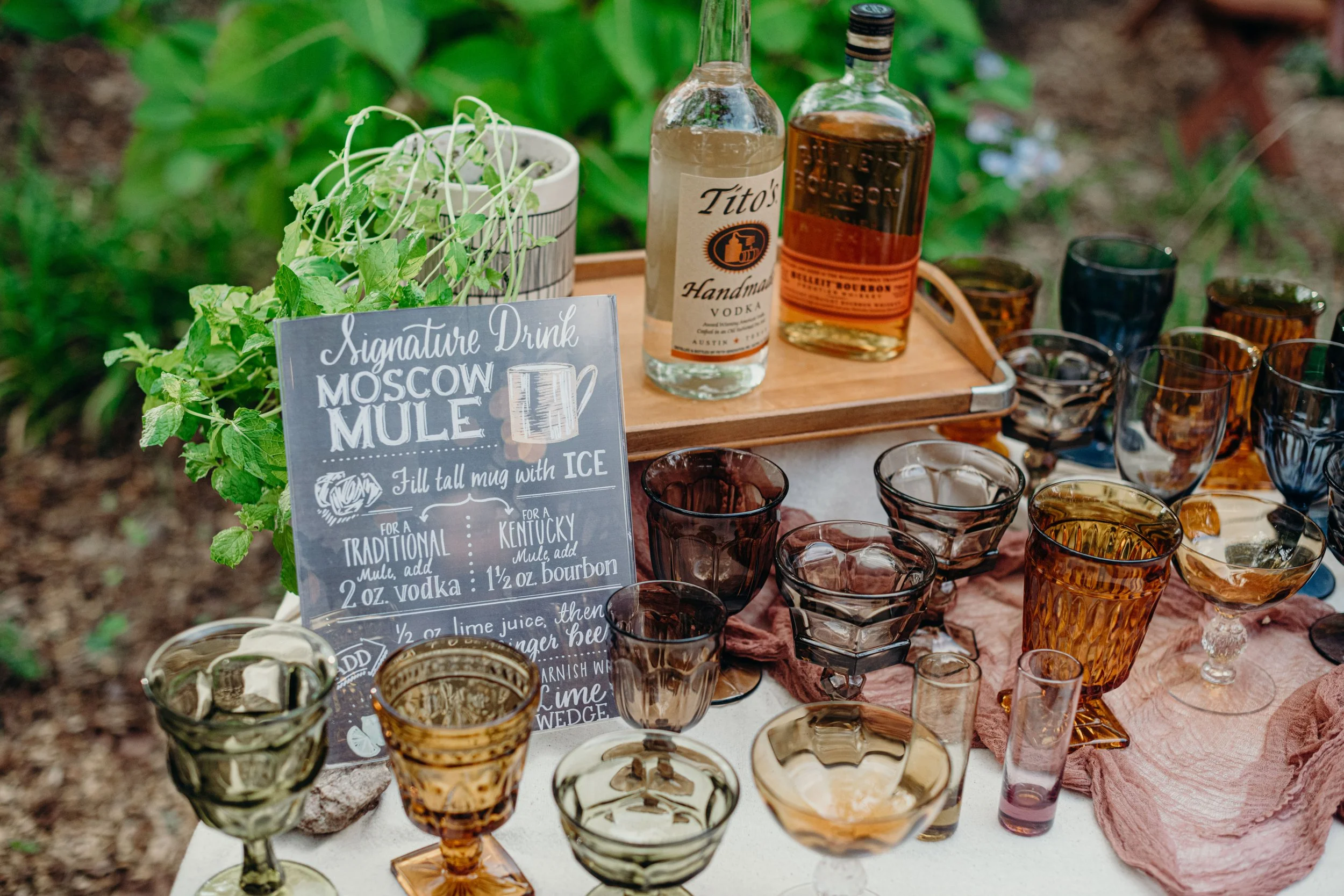 A table displaying a variety of vintage glassware, a potted mint plant, and a chalkboard sign advertising Moscow Mule drinks, outdoors surrounded by greenery.
