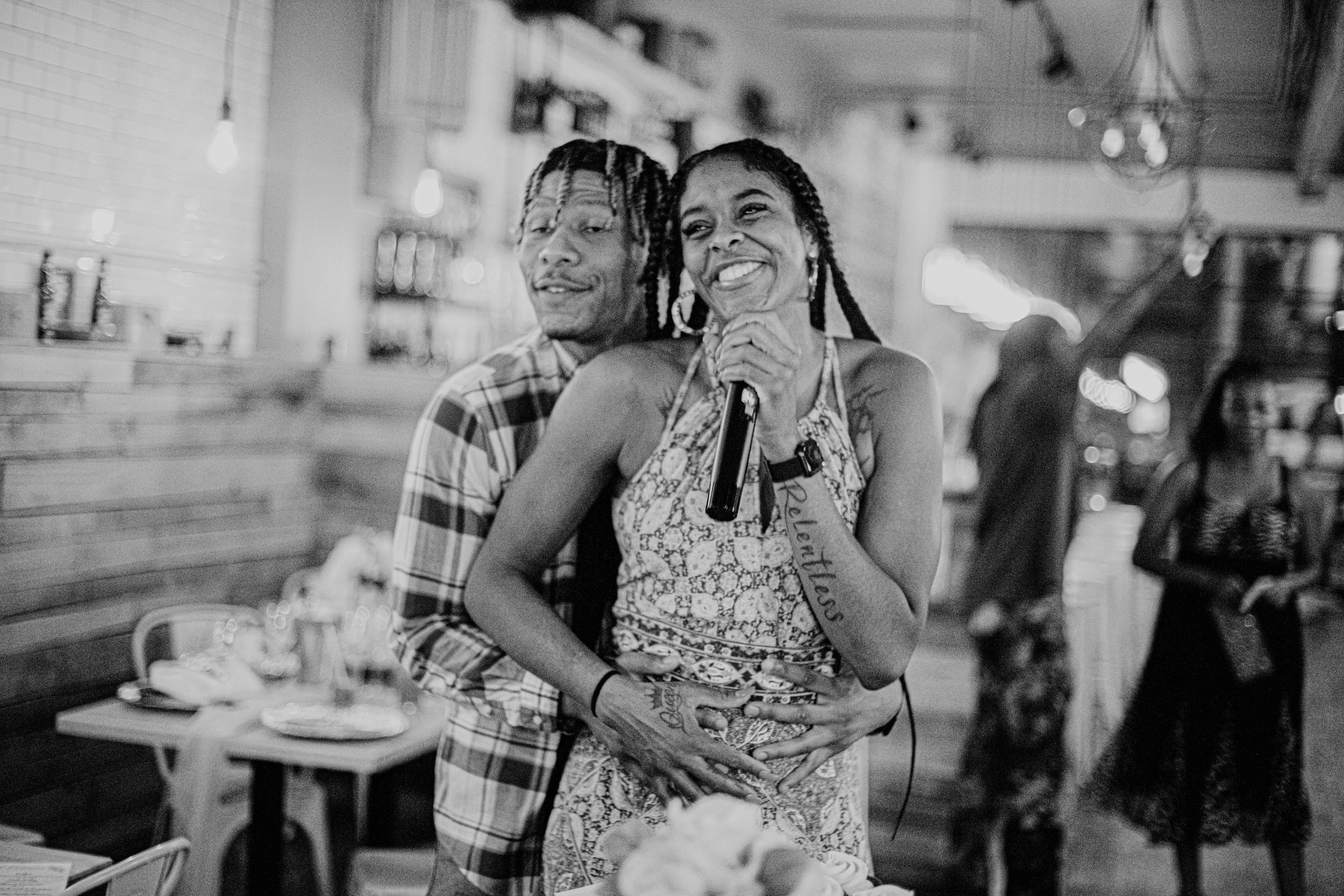 A joyful African American woman smiling while holding a microphone, with an African American man standing behind her, both in a cozy indoor setting with blurred background lighting and tables.