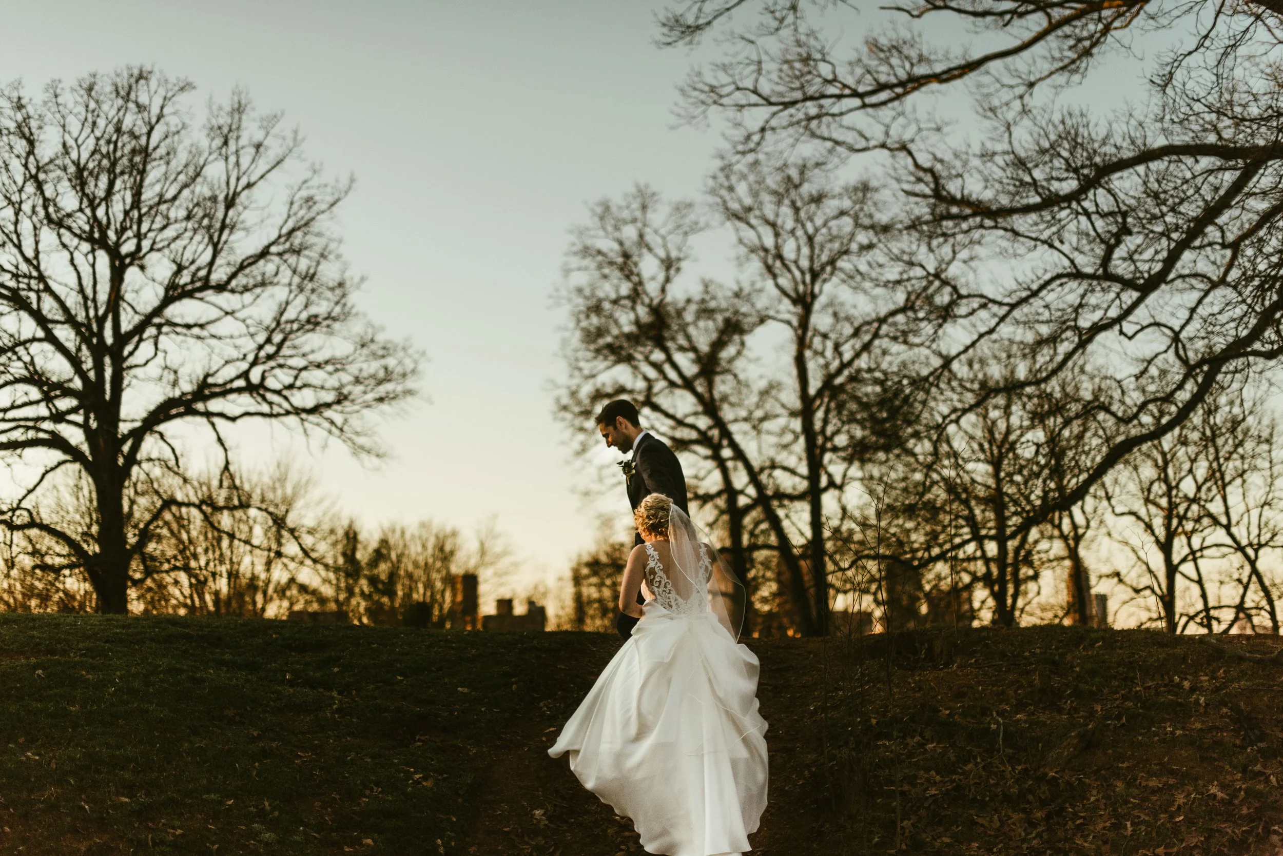 A bride and groom walking outdoors during sunset, surrounded by leafless trees. The bride wears a white wedding dress with lace details and a veil, and the groom wears a dark suit.
