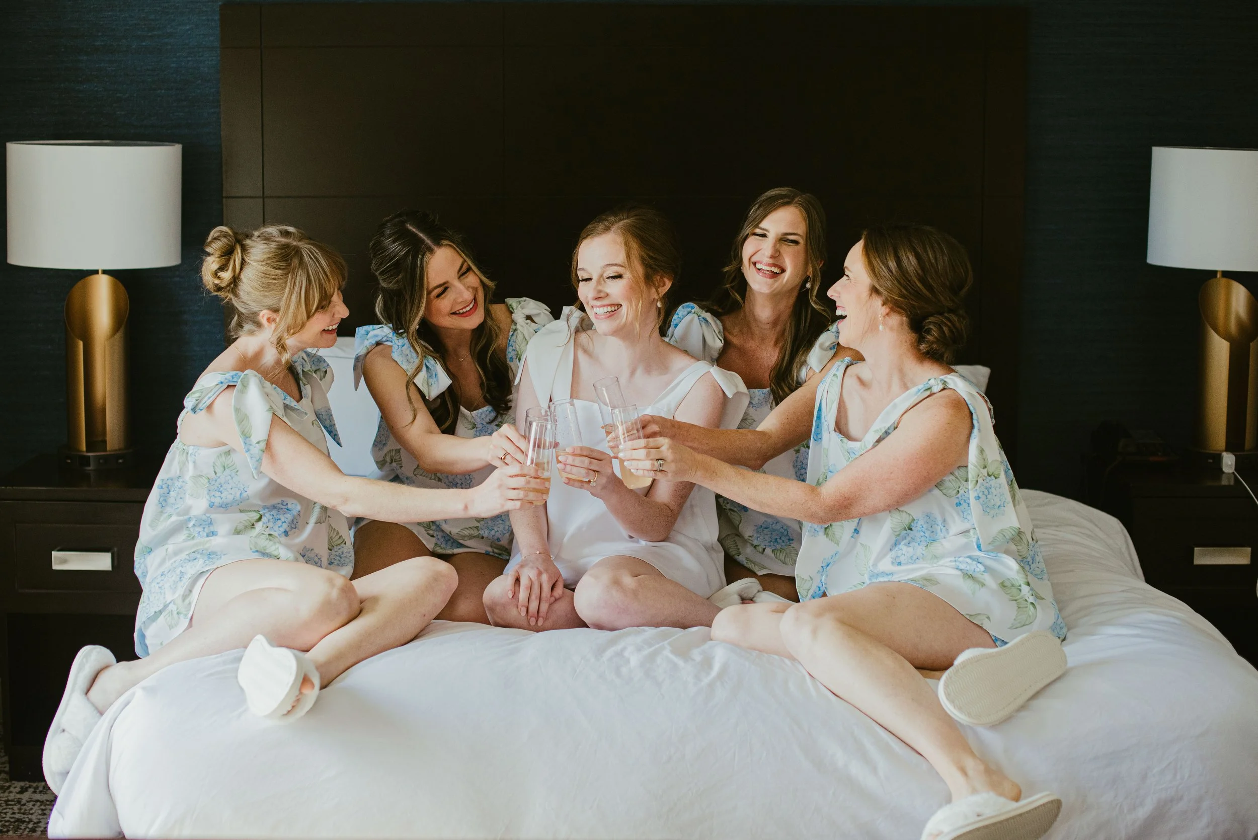 Six women sitting on a bed, celebrating and holding glasses, with a dark nightstand and lamps on either side in a hotel room.