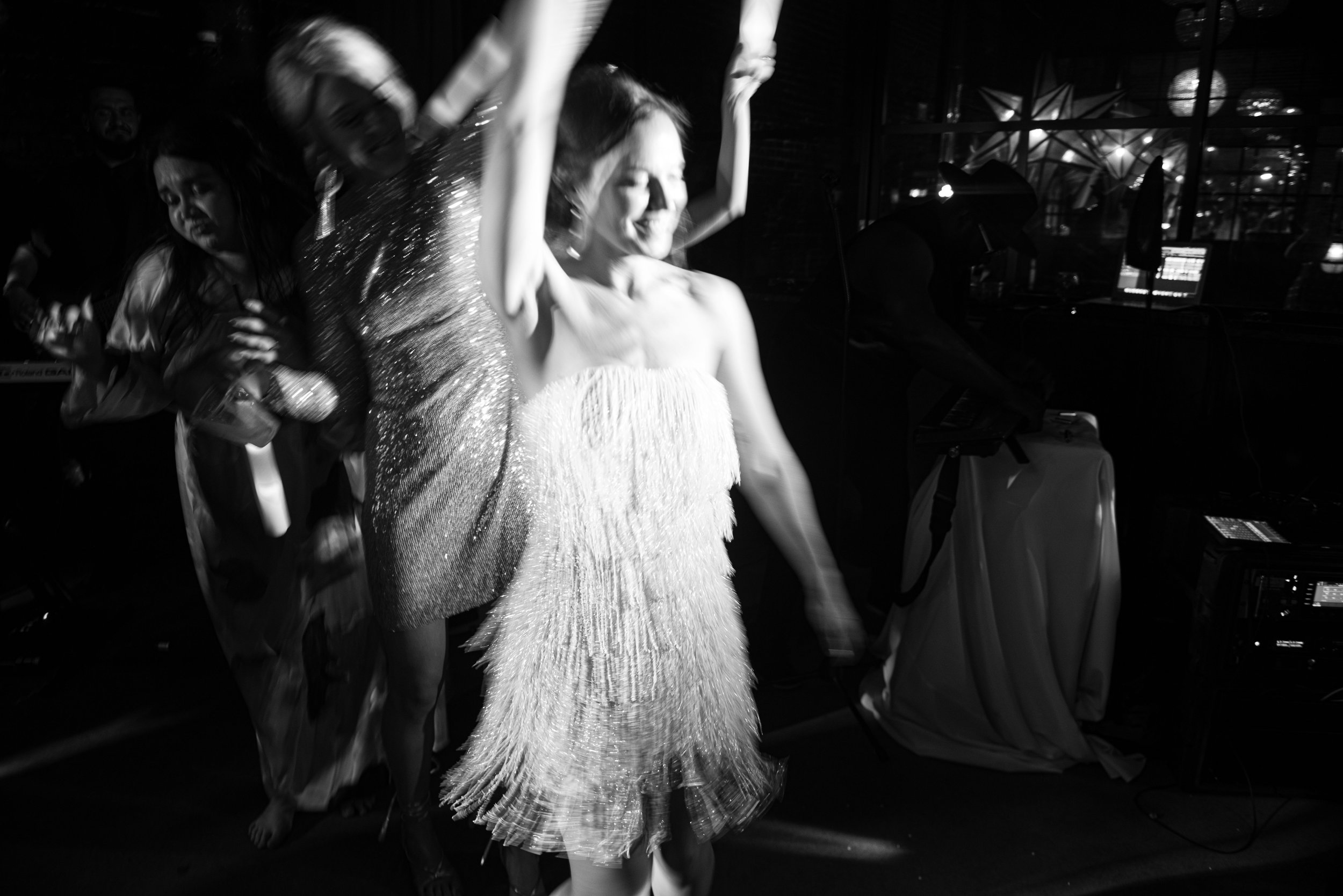 Woman dancing on stage wearing a fringed dress, with others in the background, in a dimly lit setting.