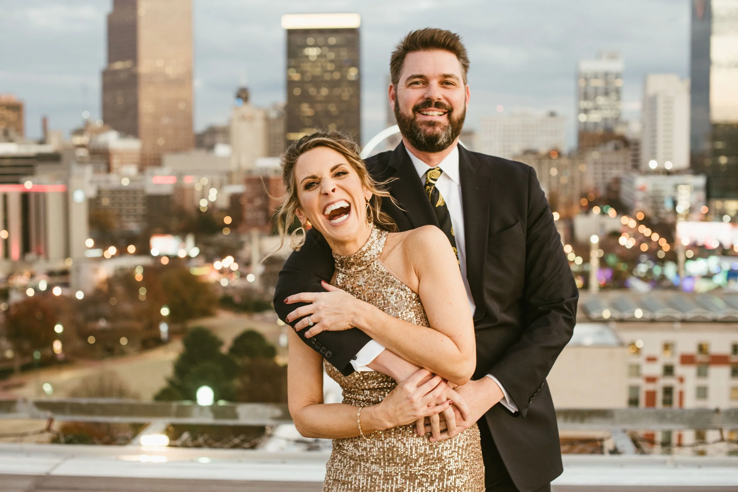 A happy couple in formal attire smiling and embracing with a city skyline in the background during twilight.