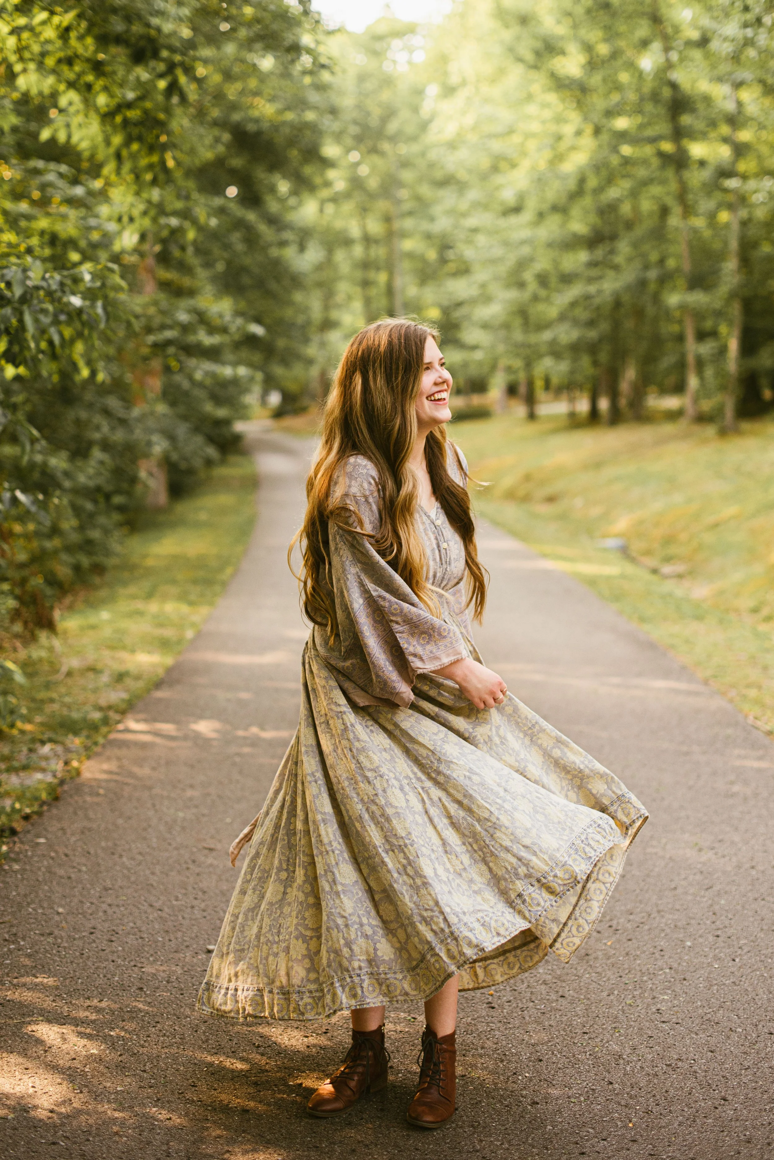 A young woman with long wavy brown hair, wearing a flowy patterned dress and brown ankle boots, is standing in the middle of a paved path surrounded by green trees and grass, smiling and looking to her left.