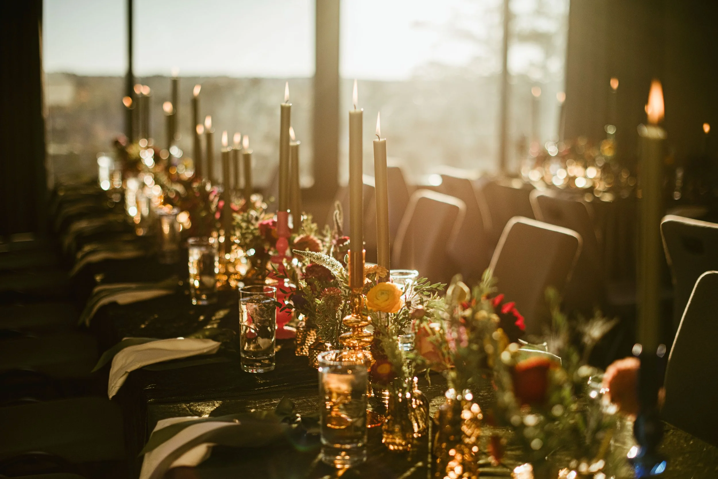 A table decorated with flowers and tall candles, with chairs around it, in a warmly lit room with large windows.