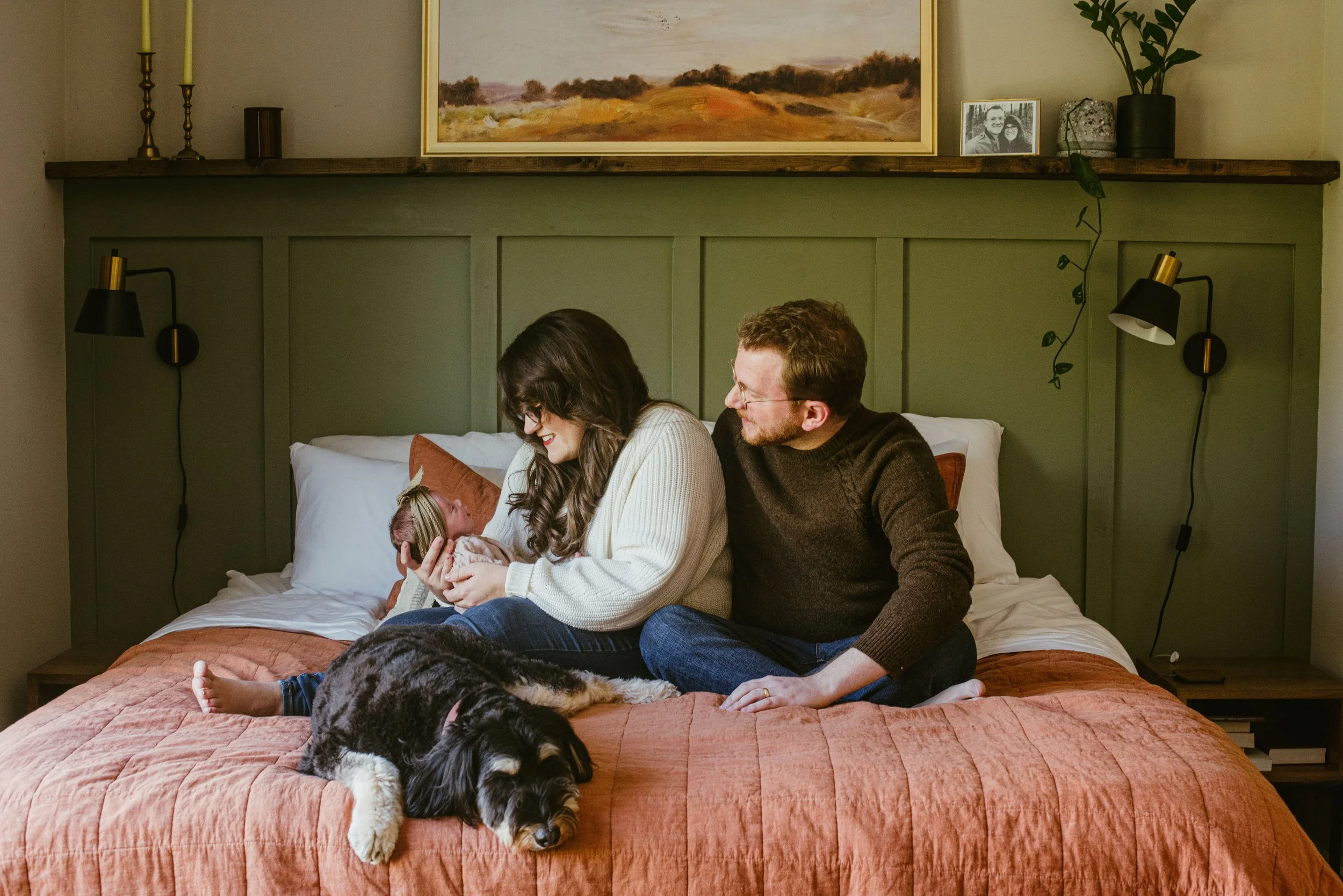 A family sitting on a bed in a bedroom, with two adults and a baby, and two dogs lying on the bed. The woman is holding the baby, and the man is smiling at the baby. The bedroom has green paneled walls, black and gold wall-mounted lamps, a wooden she