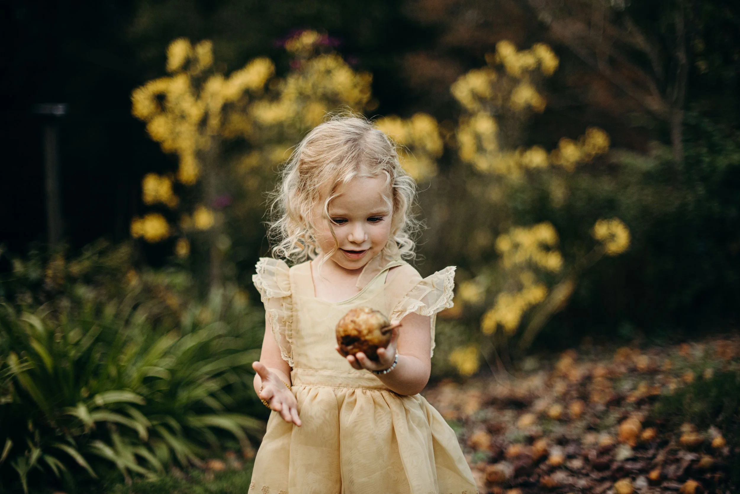 A young girl with curly blonde hair wearing a yellow dress holding a large mushroom in a garden with green plants and blurred yellow flowers in the background.