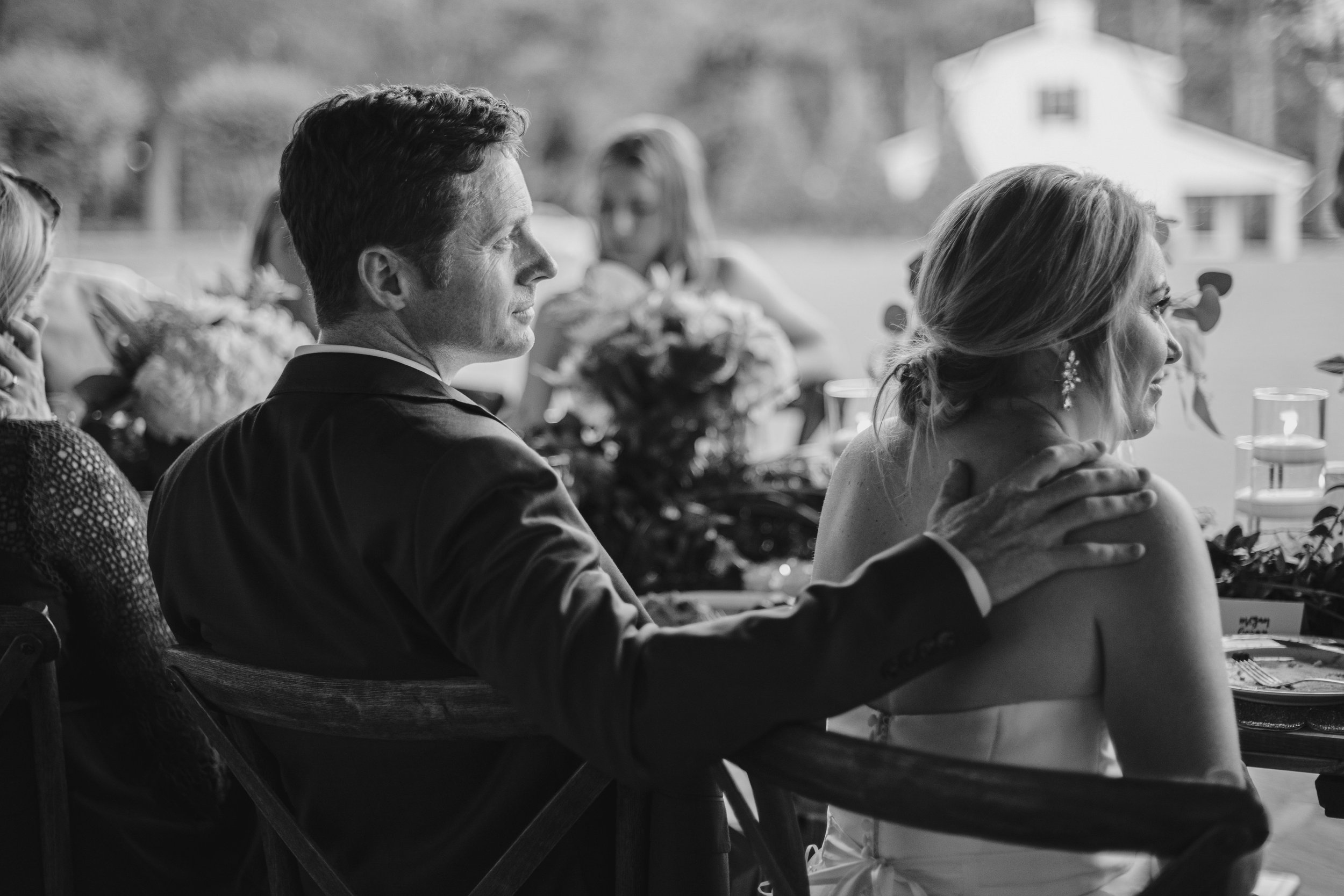 A wedding reception with guests seated at a table, including a man and a woman, both smiling and engaged with each other. The man has dark hair and is wearing a dark suit, while the woman has long, light-colored hair and earrings. The scene is outdoo