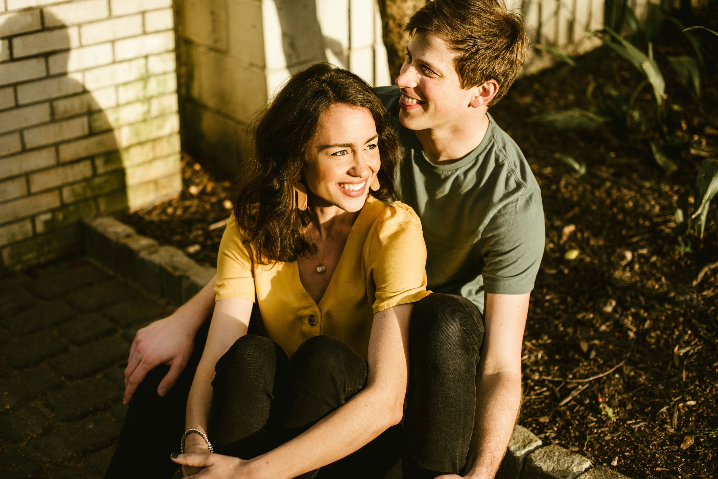 A young couple sitting outdoors and smiling at each other in warm sunlight, with a brick wall and greenery in the background.