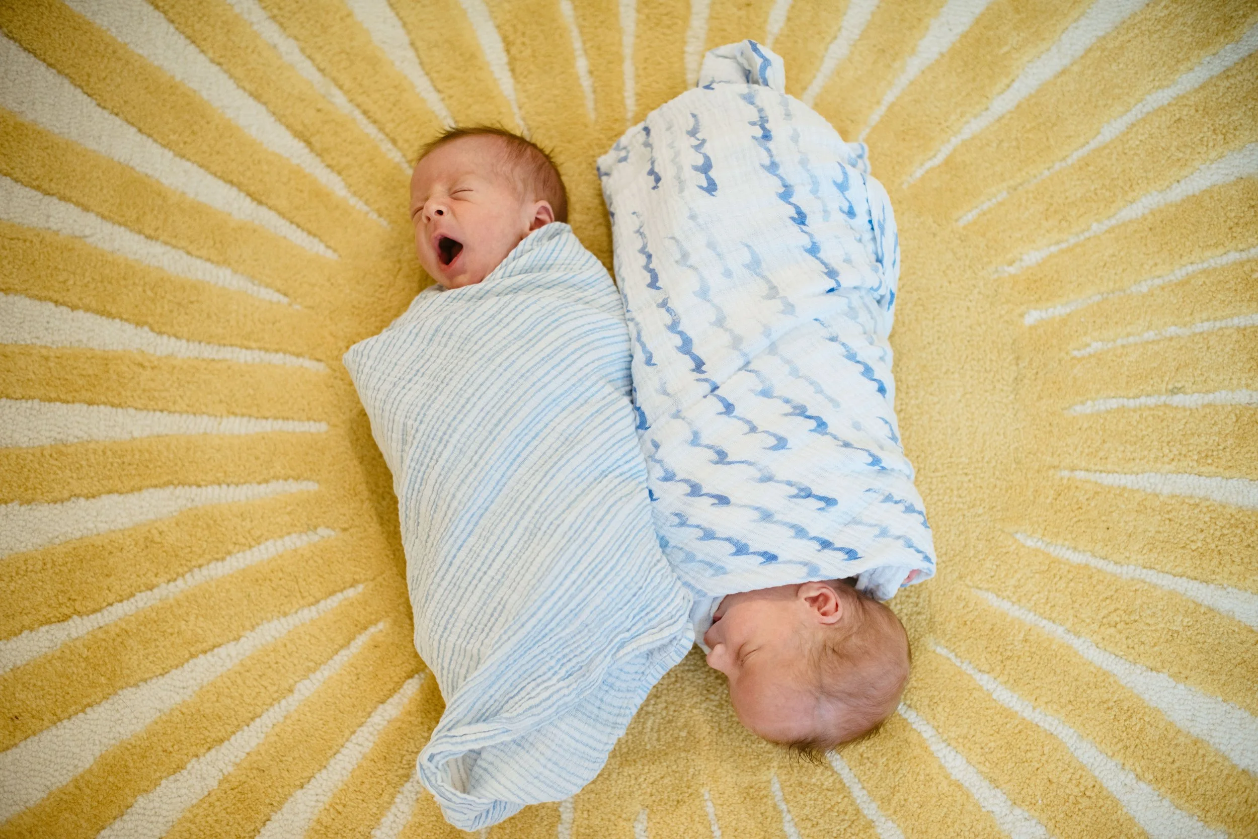 A newborn baby with red hair, wrapped in a blue and white blanket, lying on a yellow striped rug, yawning with eyes closed.