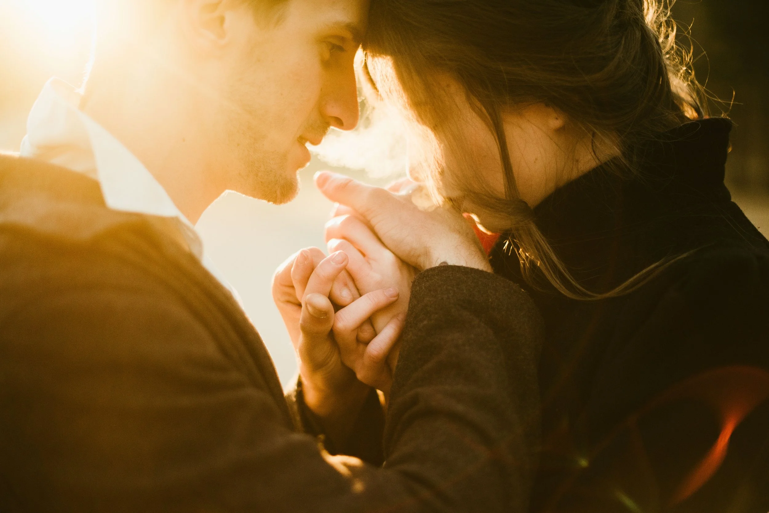 A couple sharing an intimate moment, touching foreheads and holding hands, with warm backlit lighting creating a romantic atmosphere.
