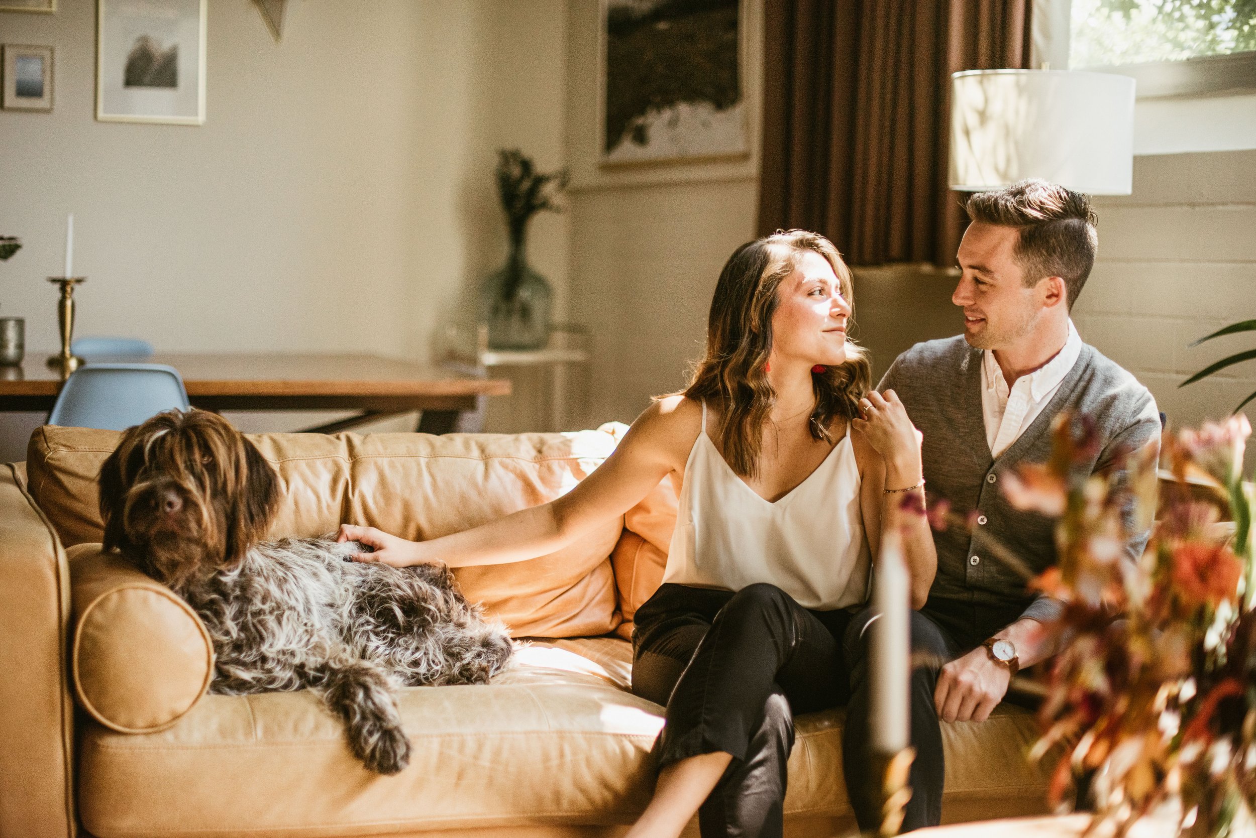 A couple sitting on a sofa in a cozy living room, smiling and looking at each other while their large dog lies on the sofa beside them.