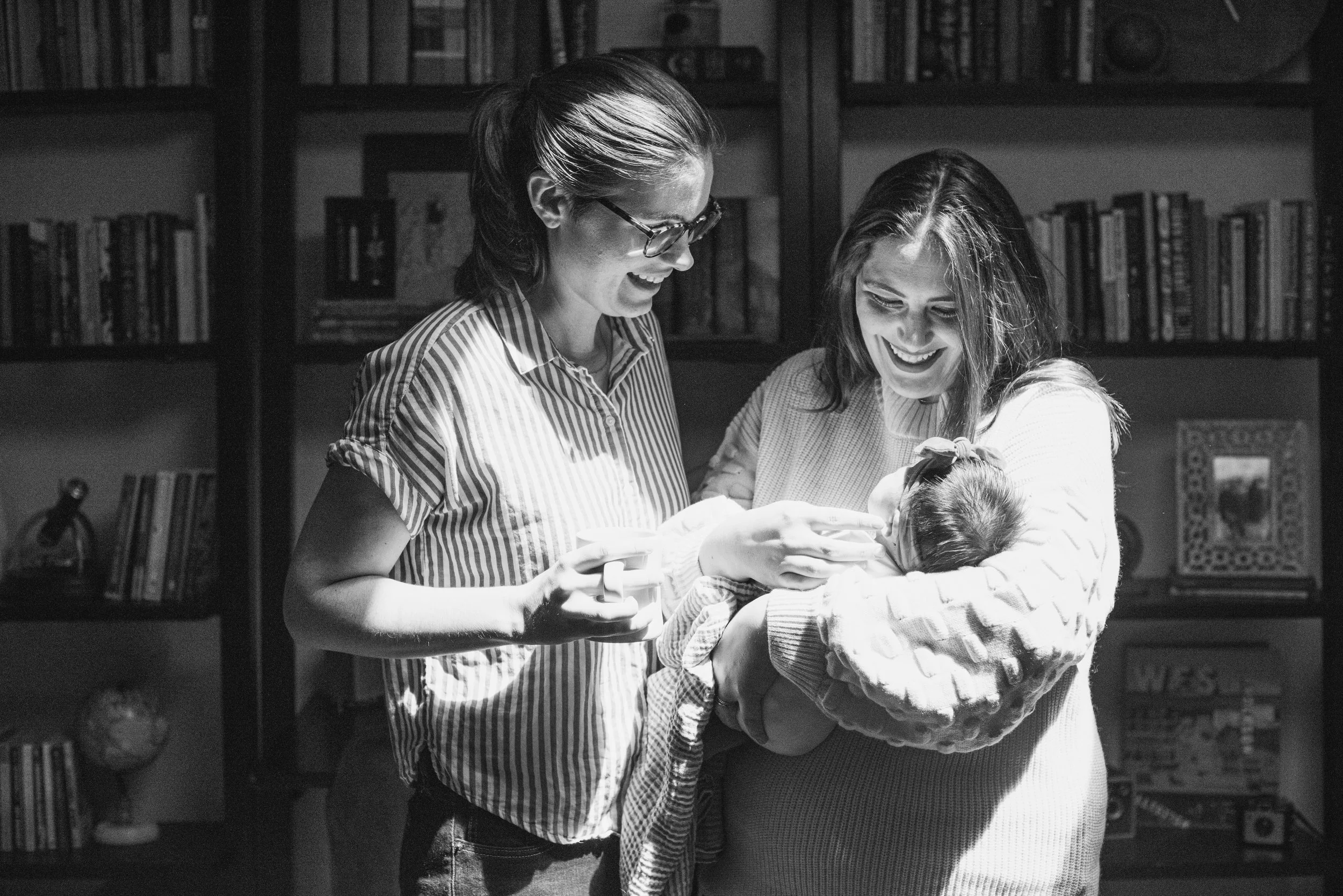 Two women are smiling and holding a baby close to them. They are inside a room with bookshelves in the background.