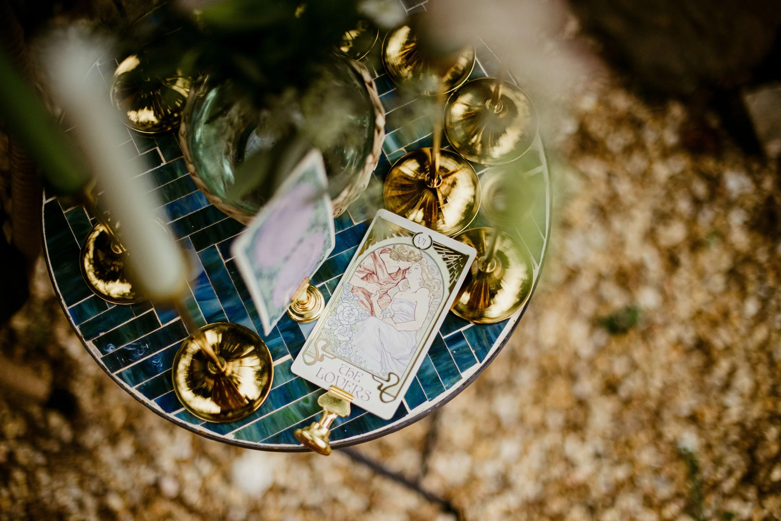 Gold-colored candle holders on a mosaic table with a card and greenery; blurred outdoor background.