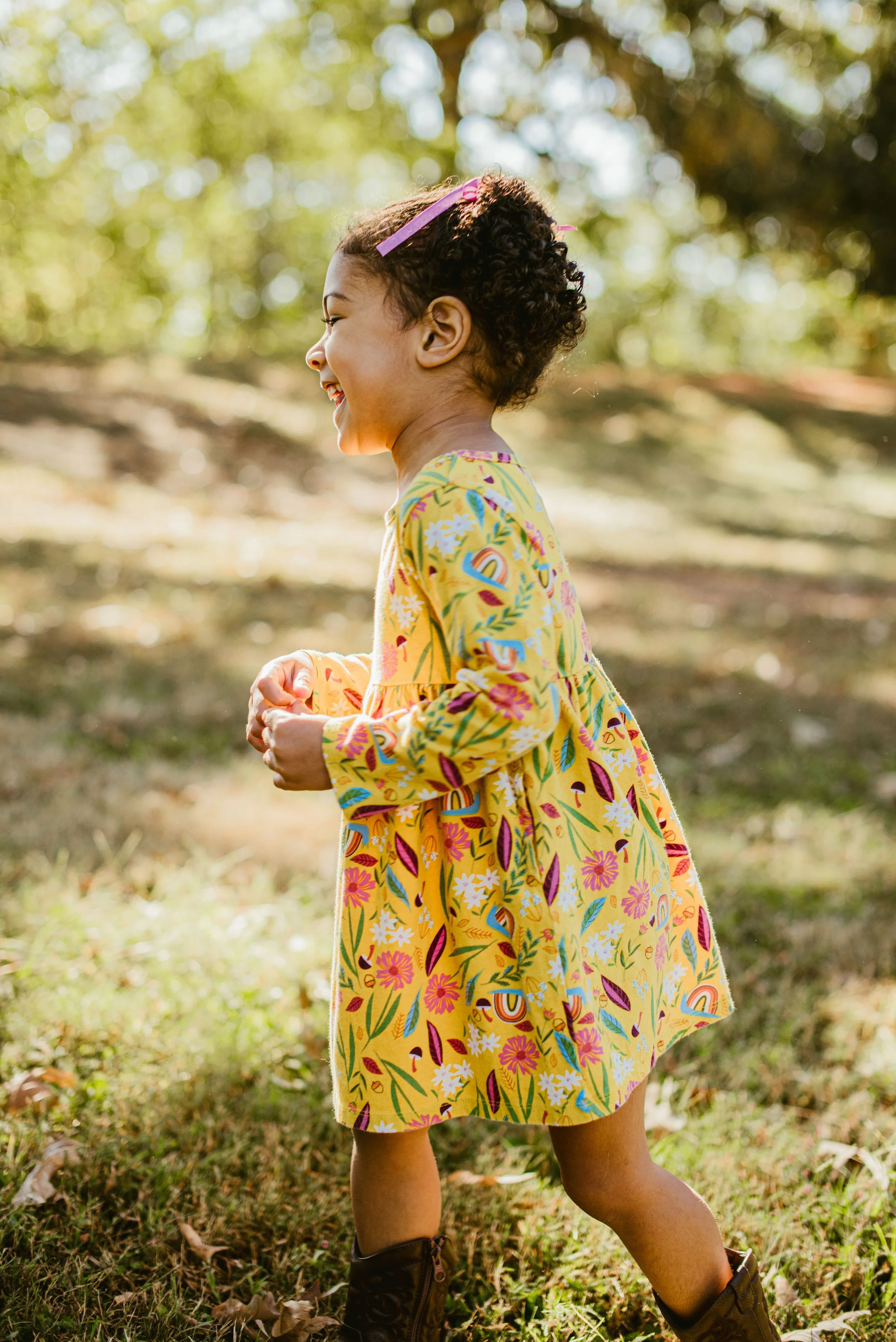 A young girl wearing a yellow floral dress and cowboy boots, smiling and walking outdoors among trees with sunlight filtering through the leaves.