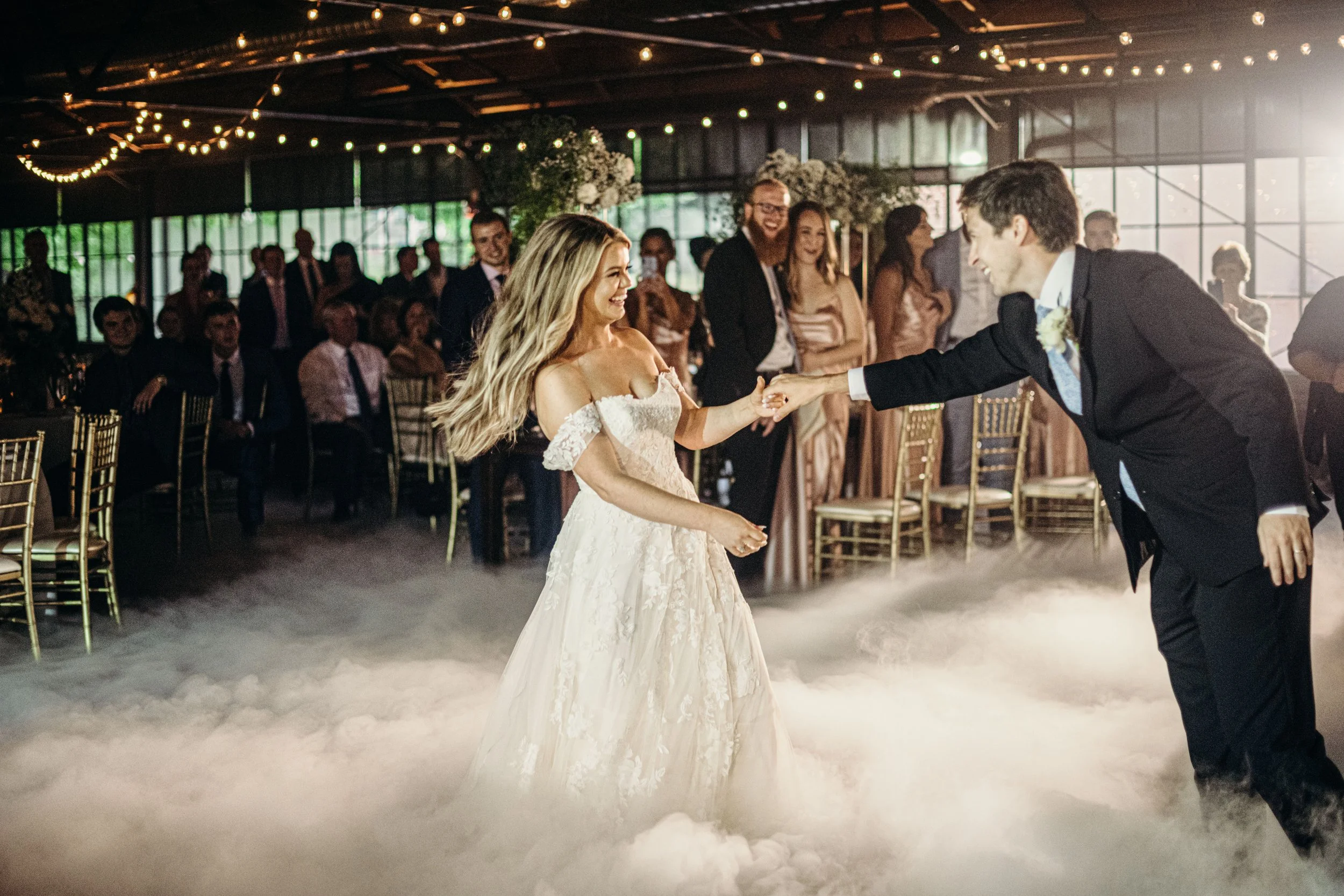 A bride and groom dancing at a wedding reception with guests seated around them and string lights overhead.