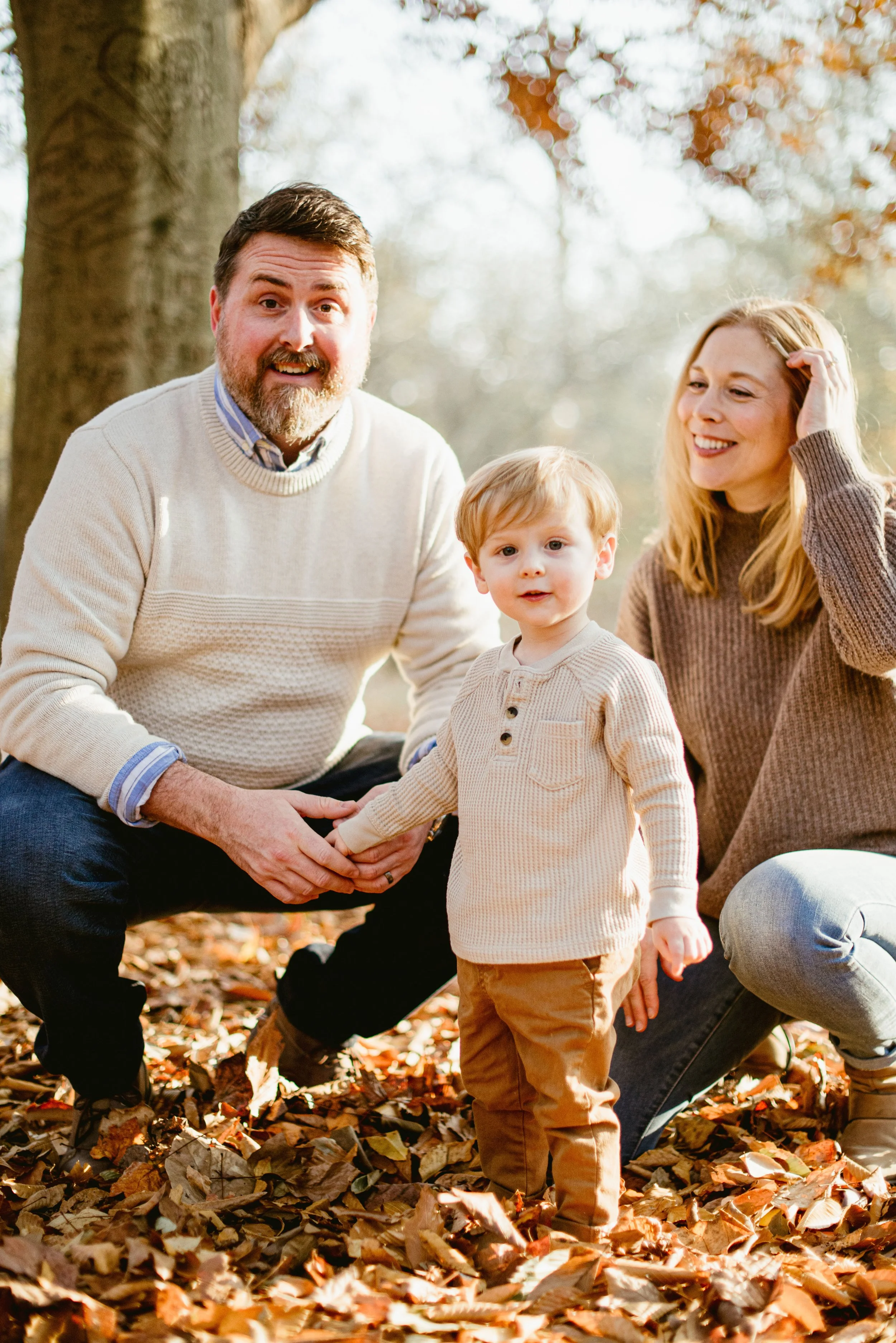 A family of three outdoors in autumn with leaves on the ground, including a man, a woman, and a young boy holding hands.