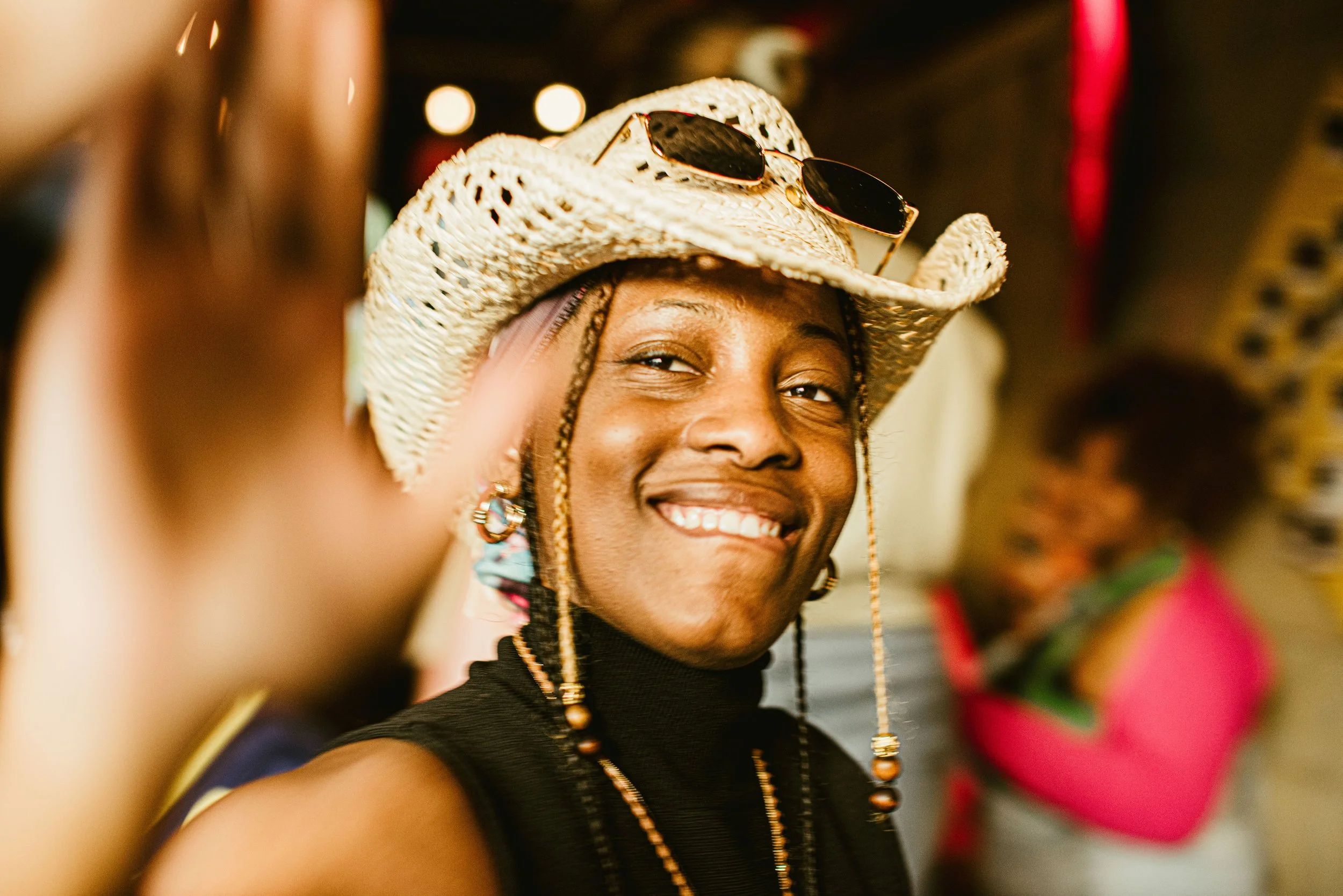 A smiling person wearing a straw hat with sunglasses on it and beaded jewelry, with other people in the background.