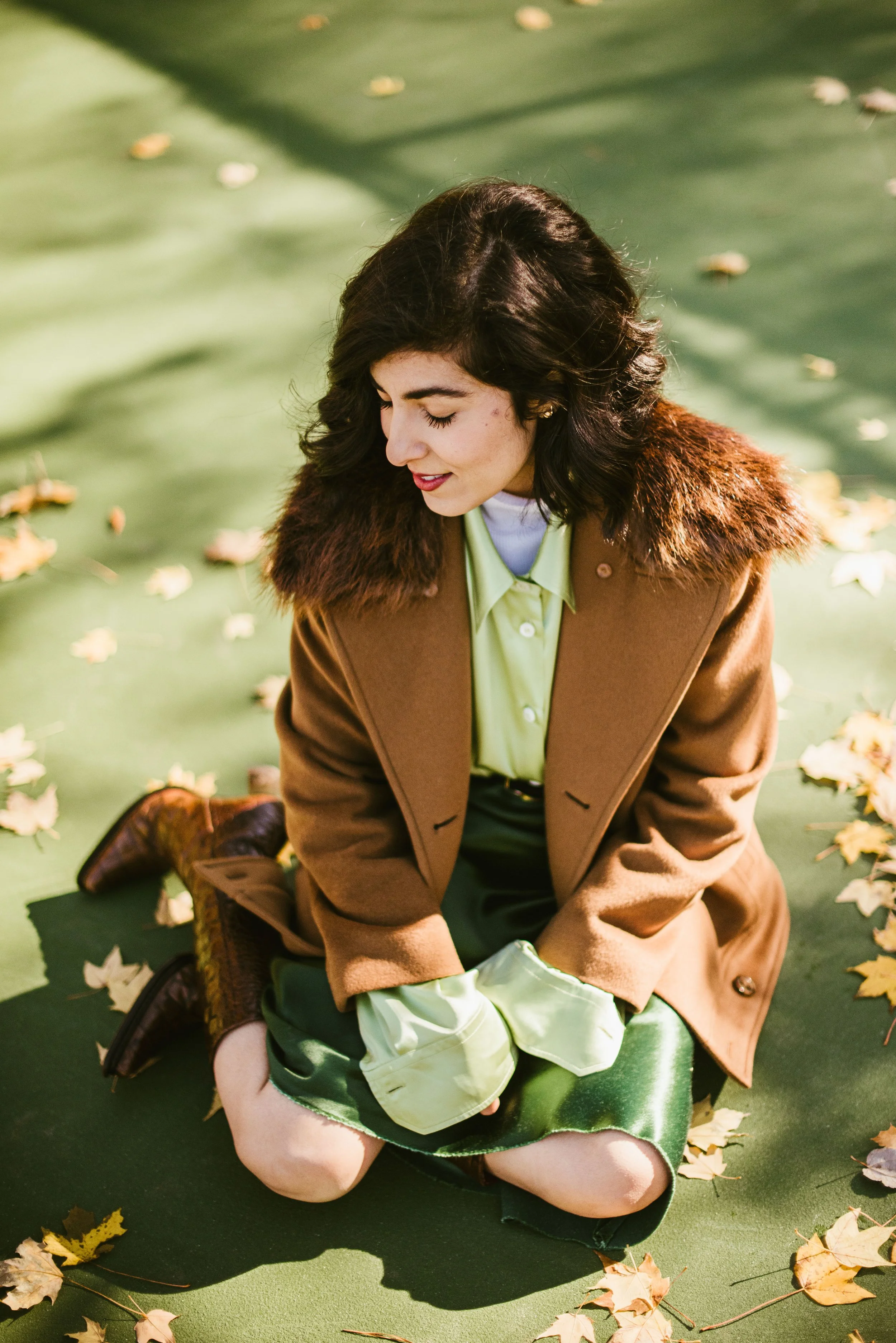 A woman with dark curly hair, wearing a brown coat with fur collar, green shirt and shorts, sitting on a green surface outdoors surrounded by fallen autumn leaves.