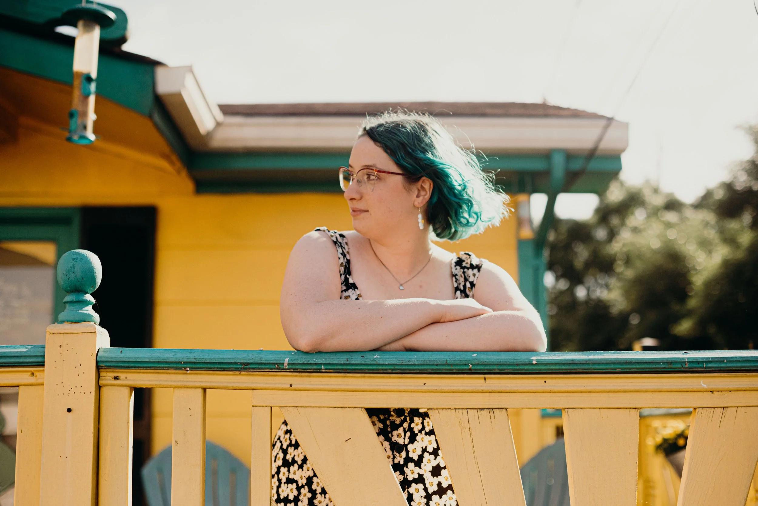 A woman with short dark green hair wearing glasses and a floral dress, leaning on a yellow wooden railing outside a colorful house with yellow, green, and brown trim, with trees in the background.