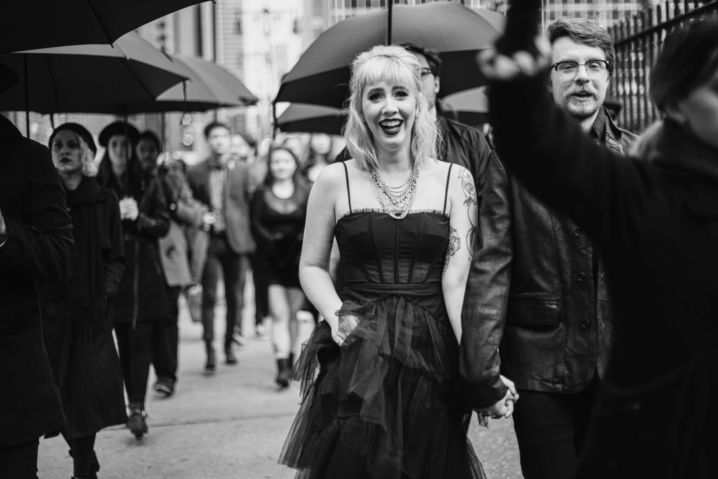 Black and white photo of a crowd of people walking in the rain on a city street, holding umbrellas. The focus is on a smiling woman with blond hair, tattoos, and wearing a dress with a tulle skirt, holding hands with a man.