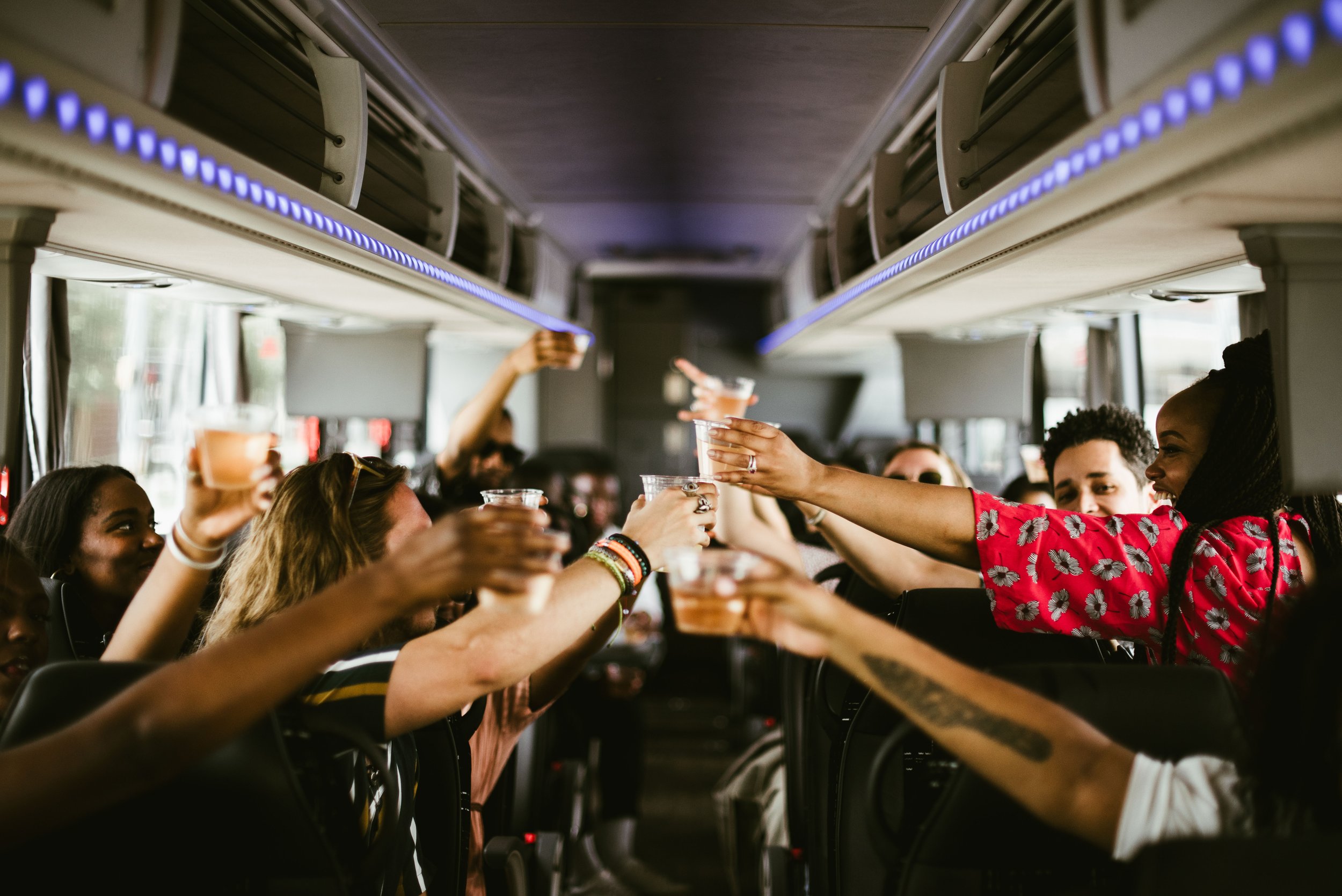 People inside a bus raising their drinks in toast, with overhead compartments and windows visible.
