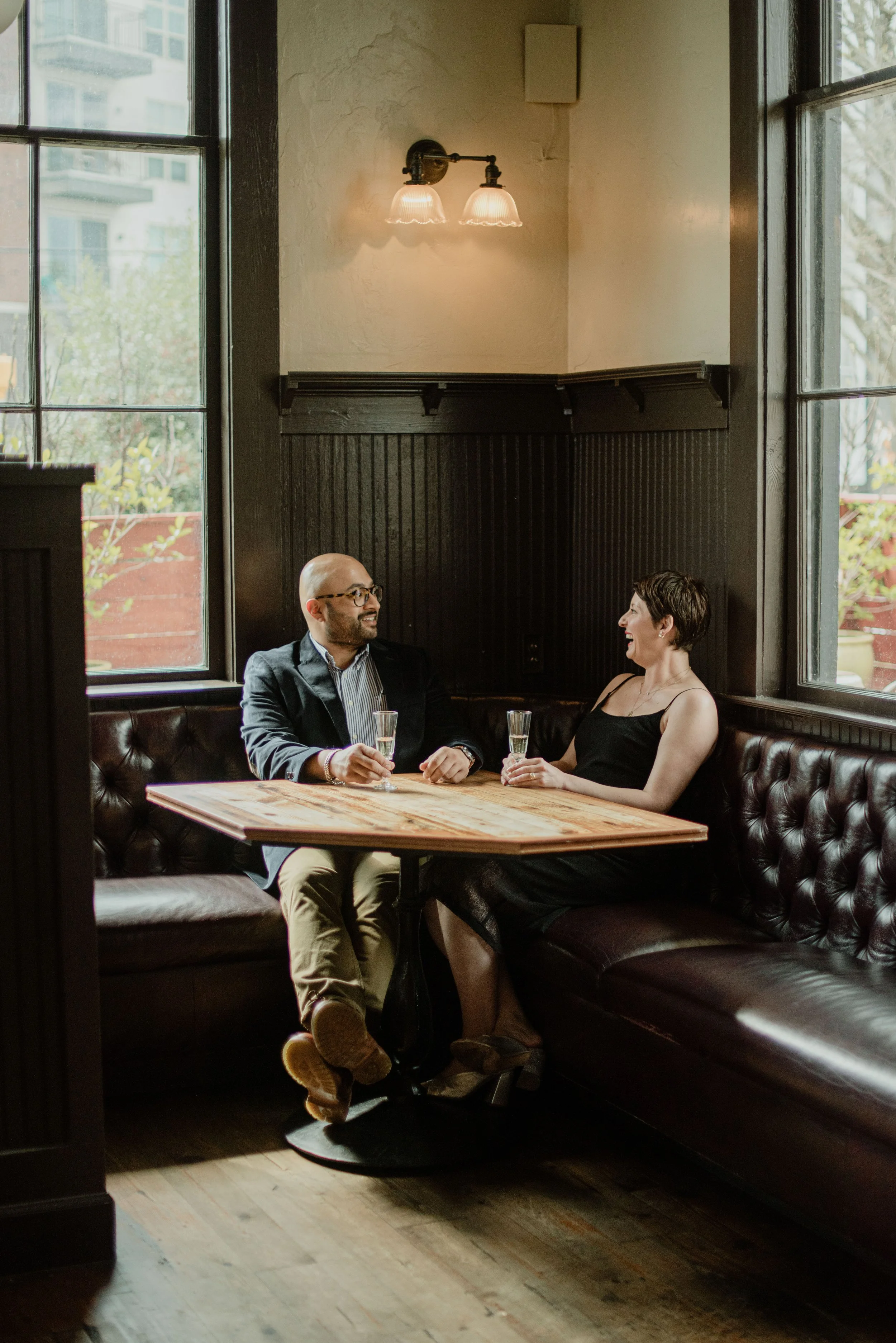 A man and a woman are sitting at a wooden table in a cozy, dimly lit restaurant or cafe, holding glasses with drinks and smiling at each other. Large windows behind them show an outdoor scene with trees and a building.