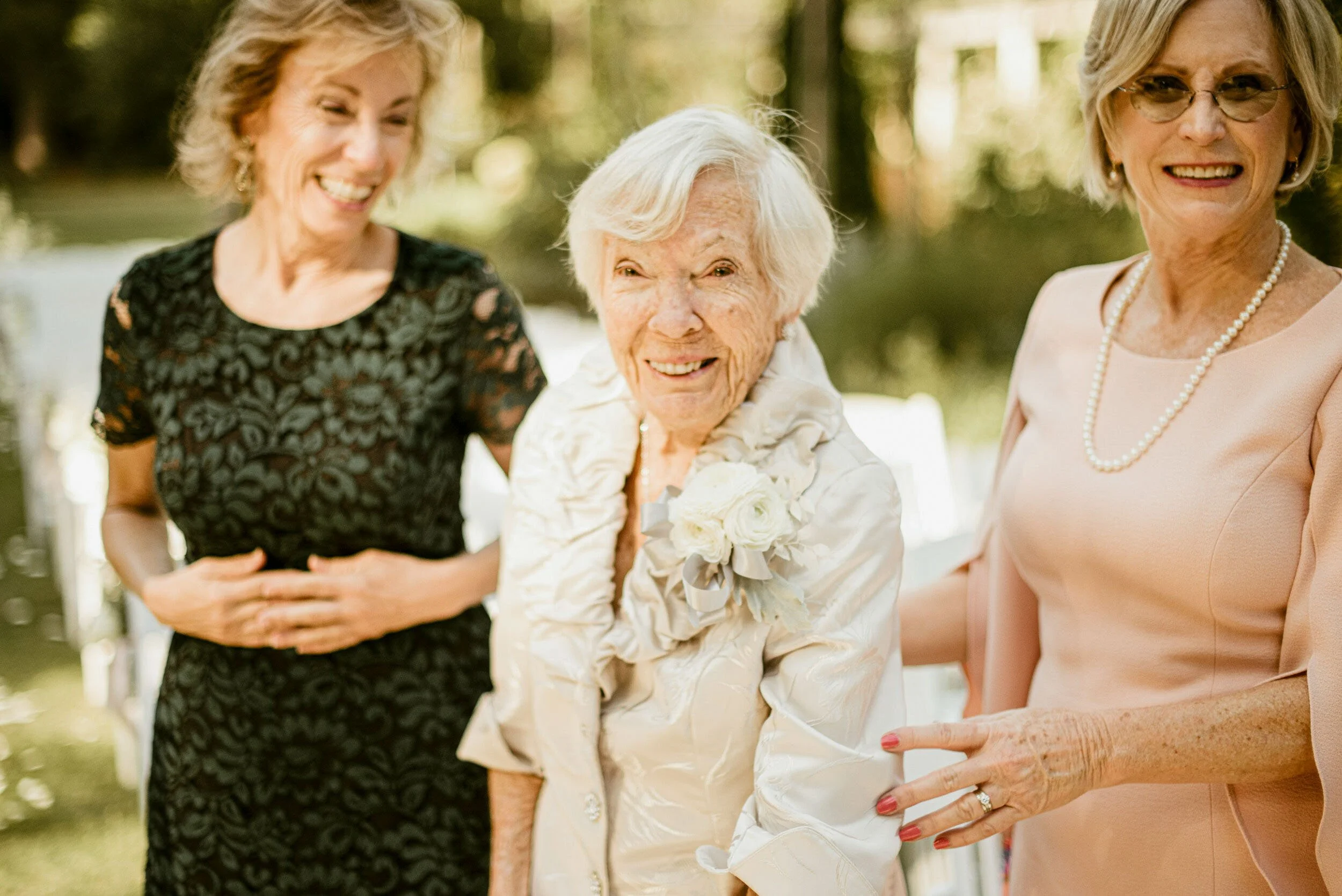 Four women, three elderly and one middle-aged, smiling and chatting outdoors in a park-like setting during daytime.