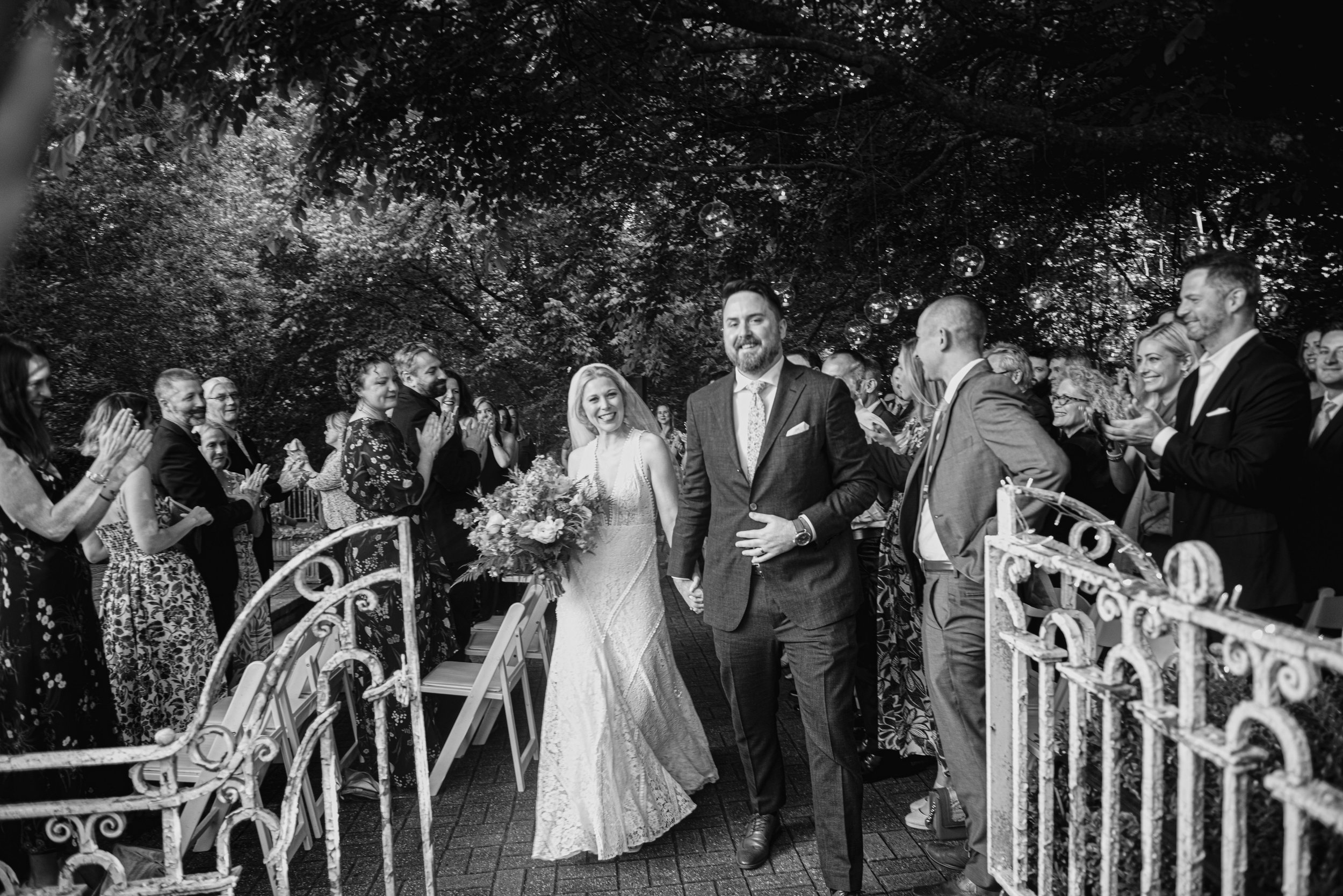 A bride and groom walking down the aisle at an outdoor wedding ceremony, surrounded by friends and family, with decorative lighting hanging above.