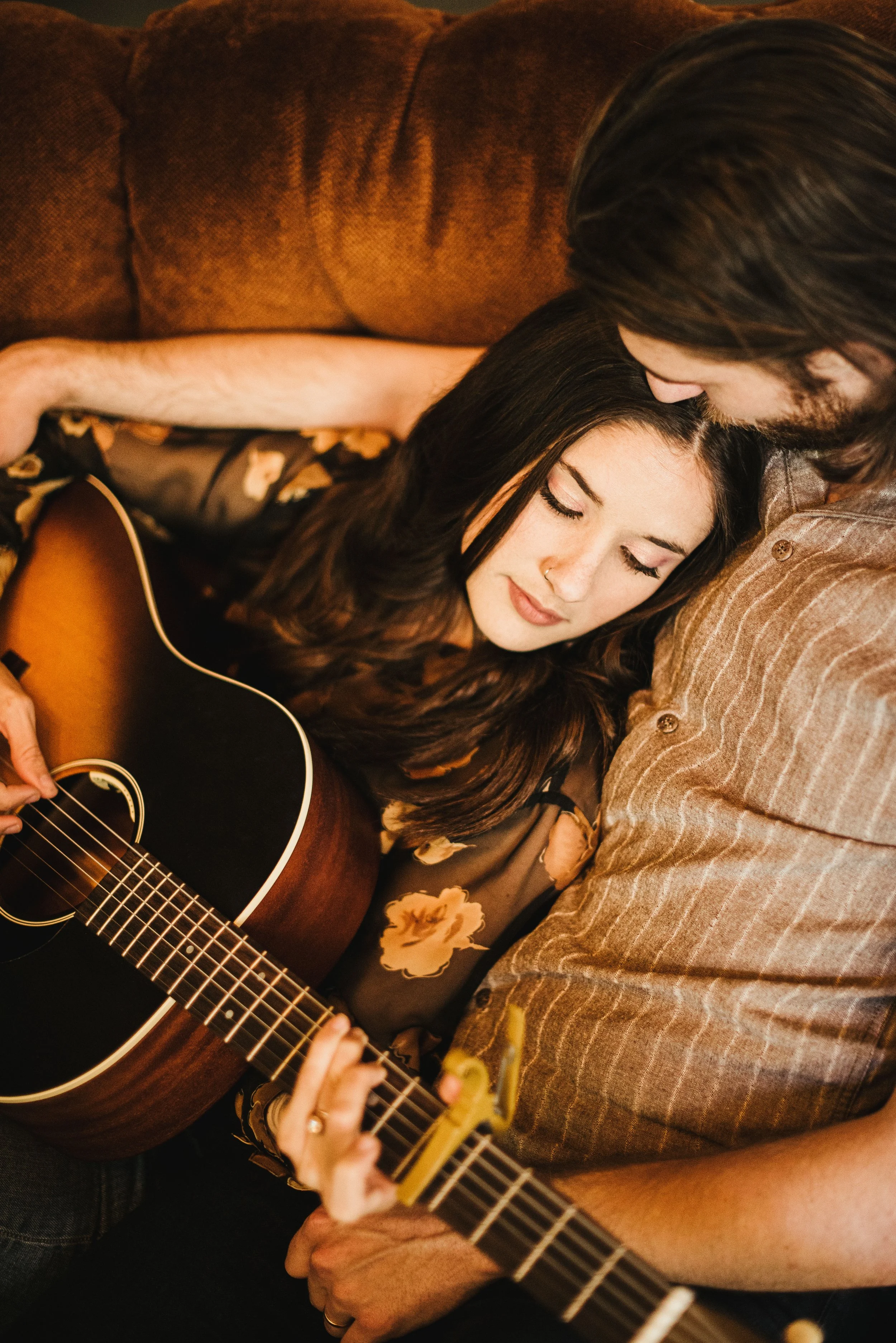 A woman with long dark hair playing an acoustic guitar, with a man with long hair and beard leaning close and kissing her forehead, both sitting on a patterned couch.