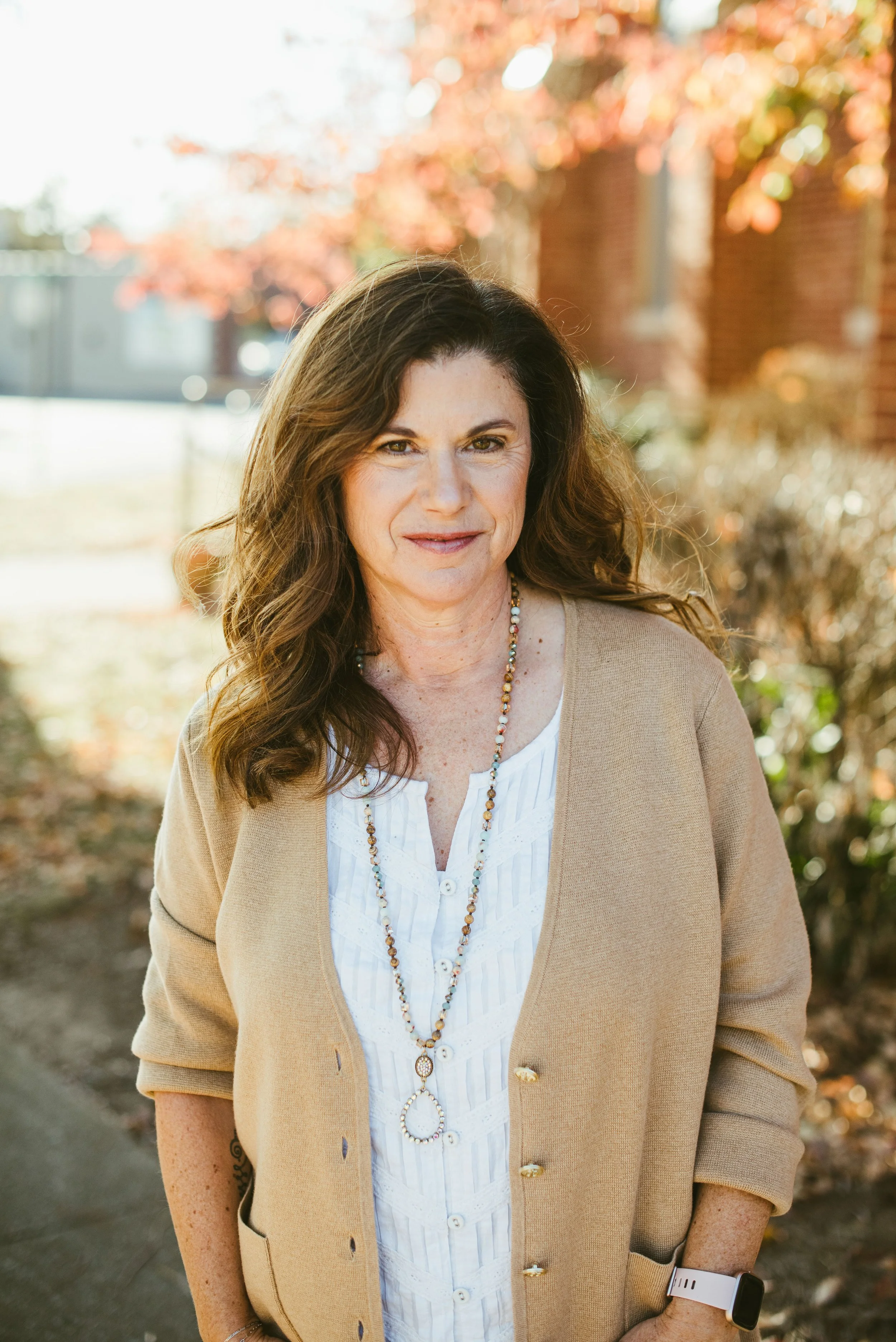 A woman with long wavy brown hair and light skin posing outdoors during autumn, wearing a beige cardigan, white blouse with buttons, a beaded necklace, and a white smartwatch on her left wrist. The background features colorful fall trees and a brick 