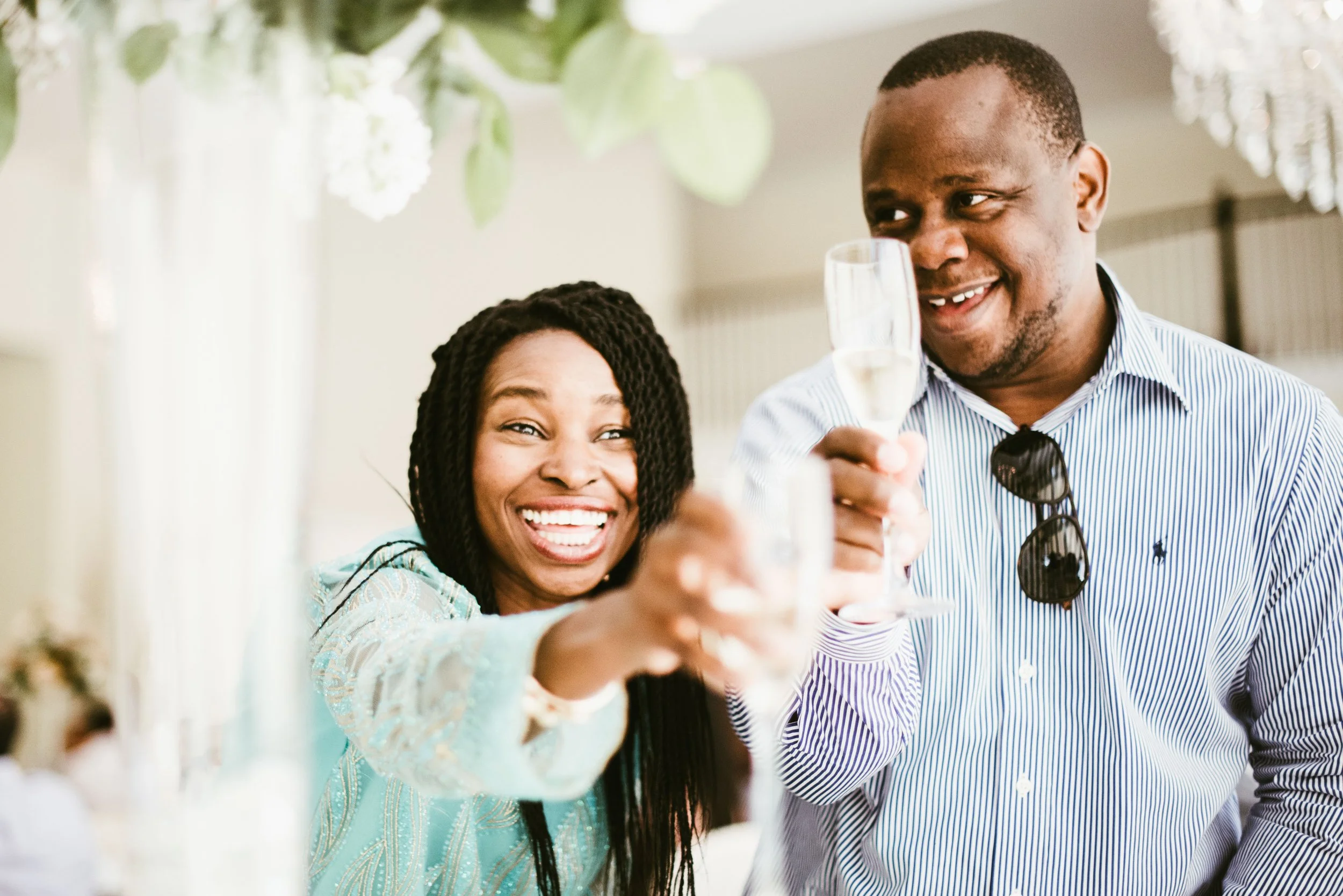 Two people smiling and taking a selfie at a gathering or celebration, with drinks in hand and festive decorations in the background.