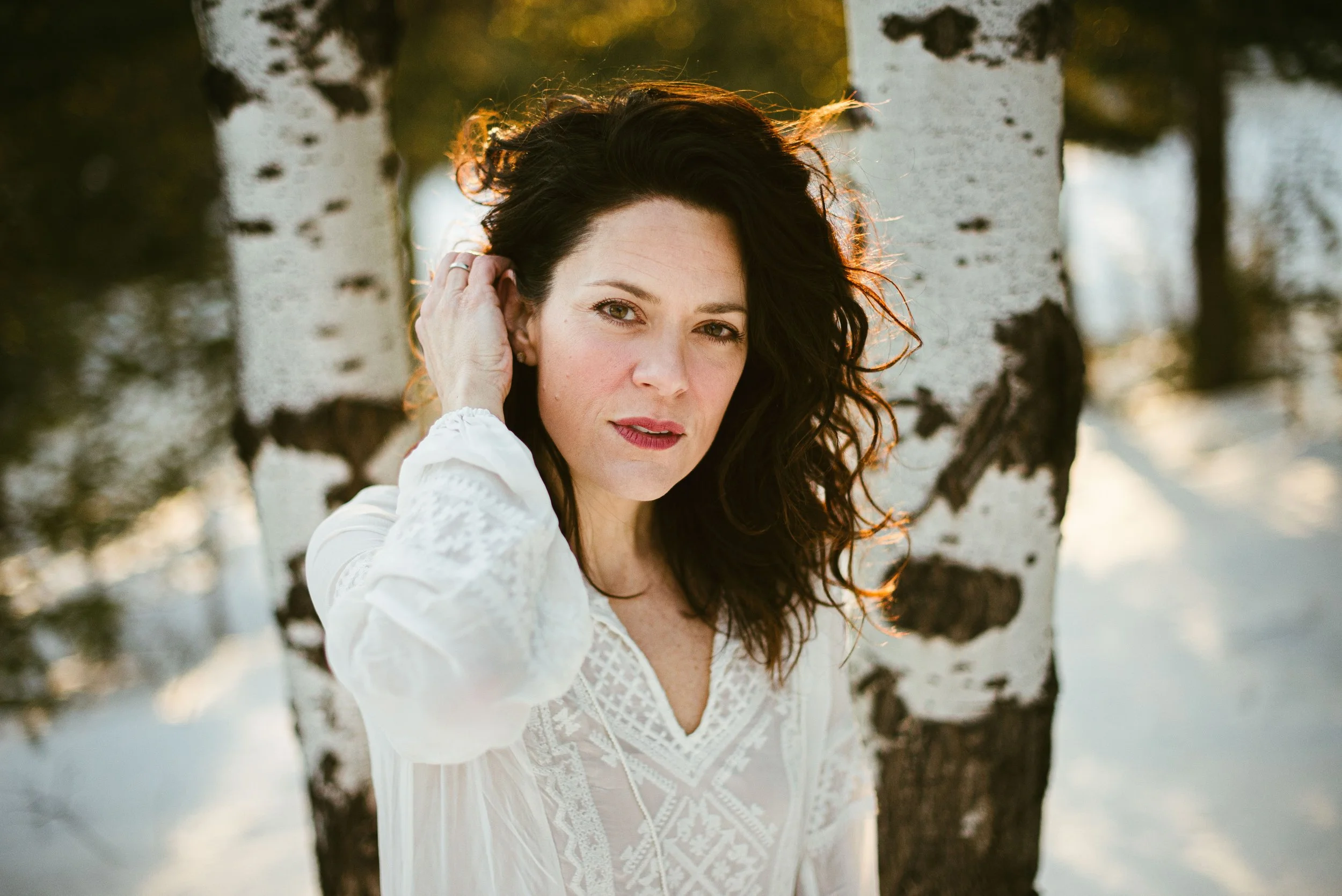 A woman with dark, curly hair and light-colored eyes standing between two white birch trees, wearing a white lace dress, with the sun setting behind her.