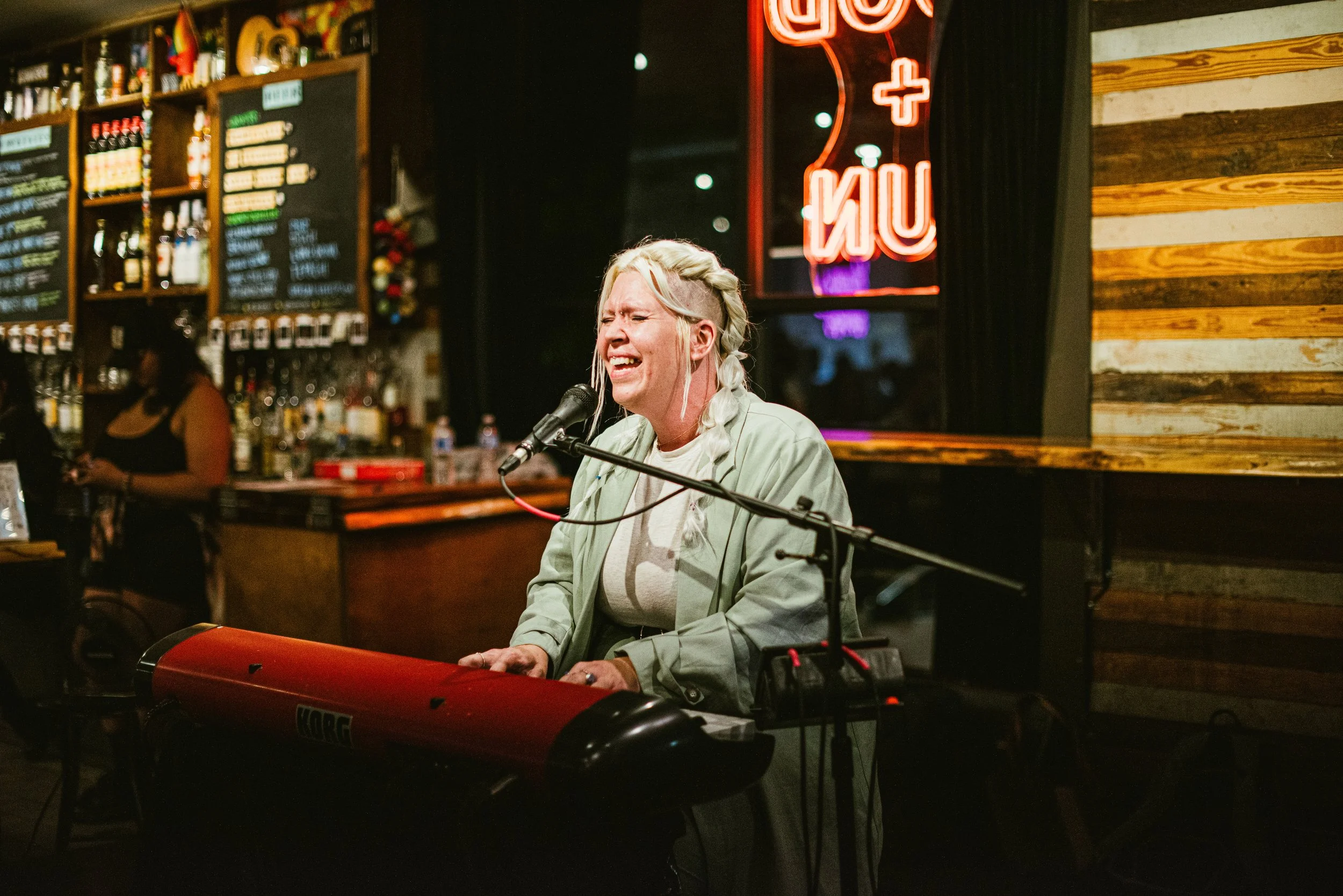 A woman with bleached hair performing with a keyboard and microphone in a bar or restaurant at night. There are chalkboard menus and a neon sign in the background.